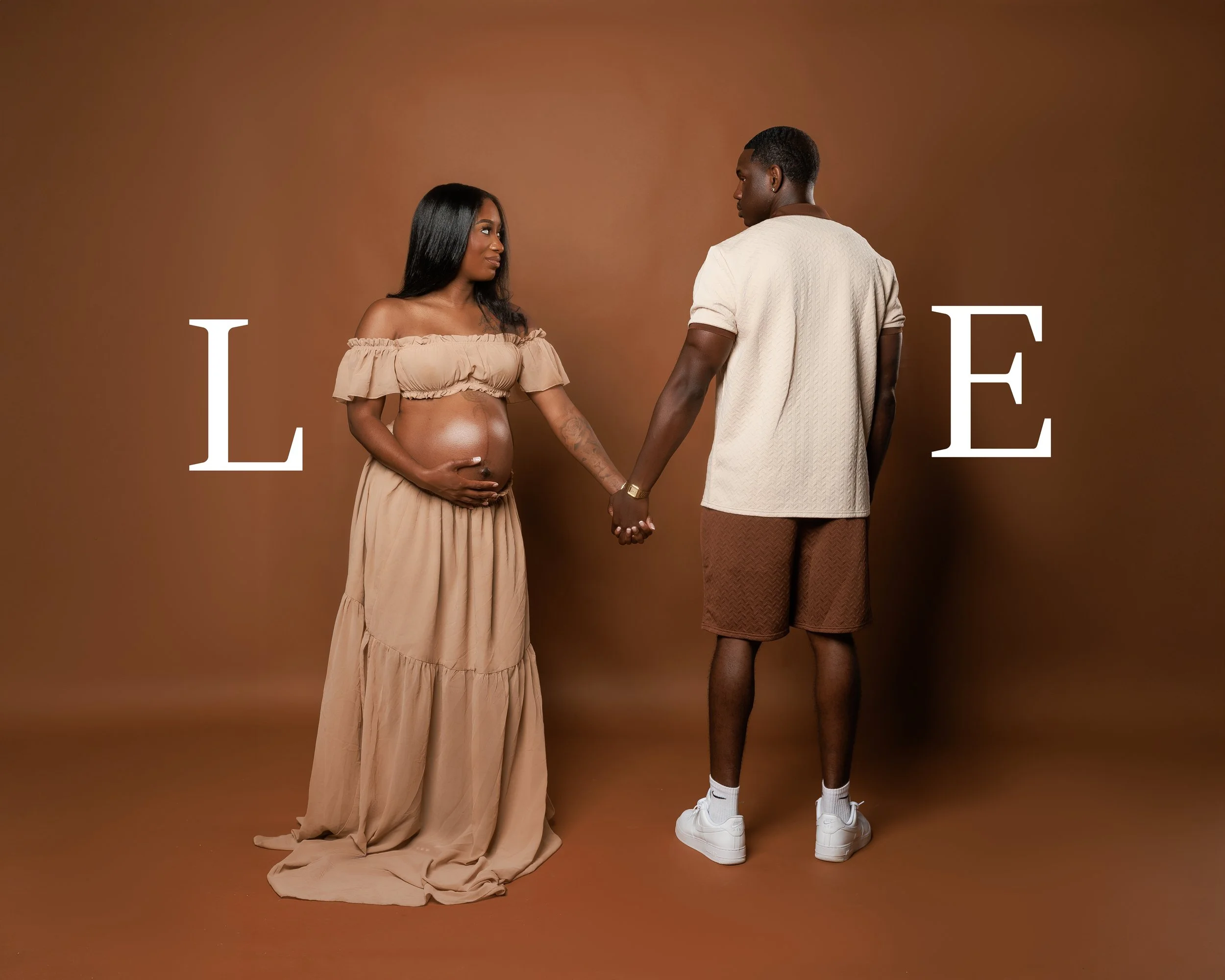 A pregnant woman with long black hair wearing a beige off-the-shoulder top and a long beige skirt holding her belly, holding hands with a young man with short black hair wearing a beige short-sleeve shirt, brown shorts, white sneakers, and black shee