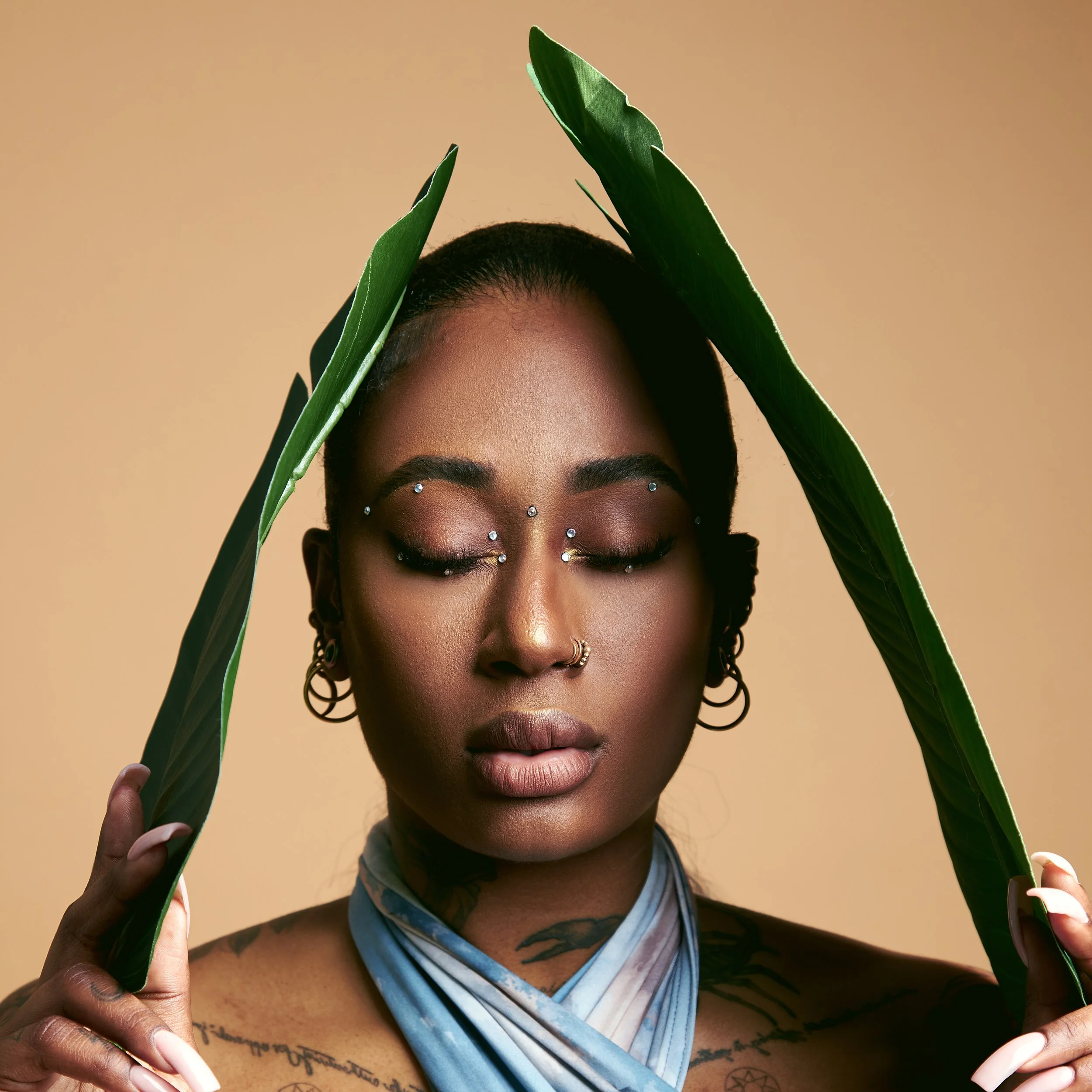 A woman with short dark hair, multiple facial piercings, earrings, and tattoos on her shoulder and chest, holding green leaves framing her face against a beige background.