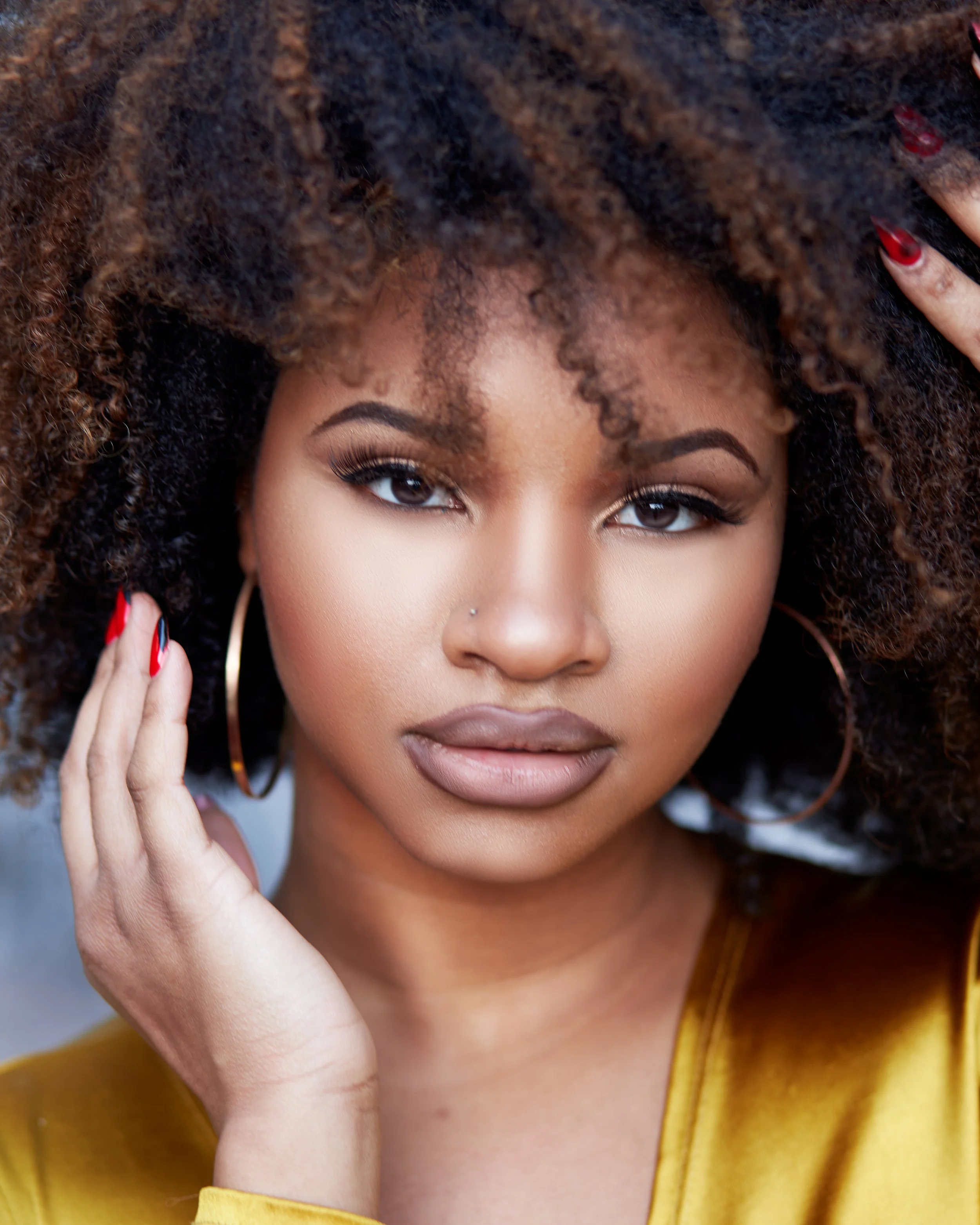 Close-up of a woman with curly hair and brown eyes, wearing makeup and a yellow top, touching her hair with her right hand, which has red painted nails and large hoop earrings.