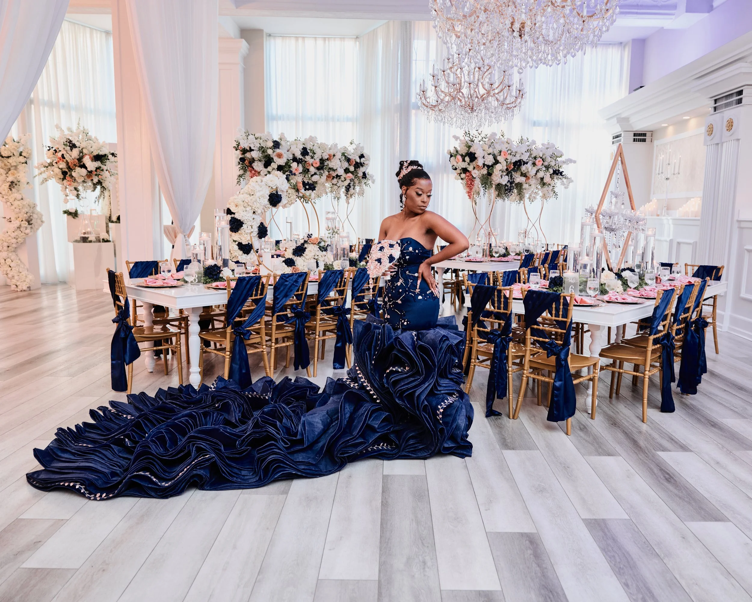 Elegant event hall decorated with white and pink floral arrangements, chandeliers, and navy blue accents on chairs and the bride's dress, featuring a long navy blue gown with floral embellishments.