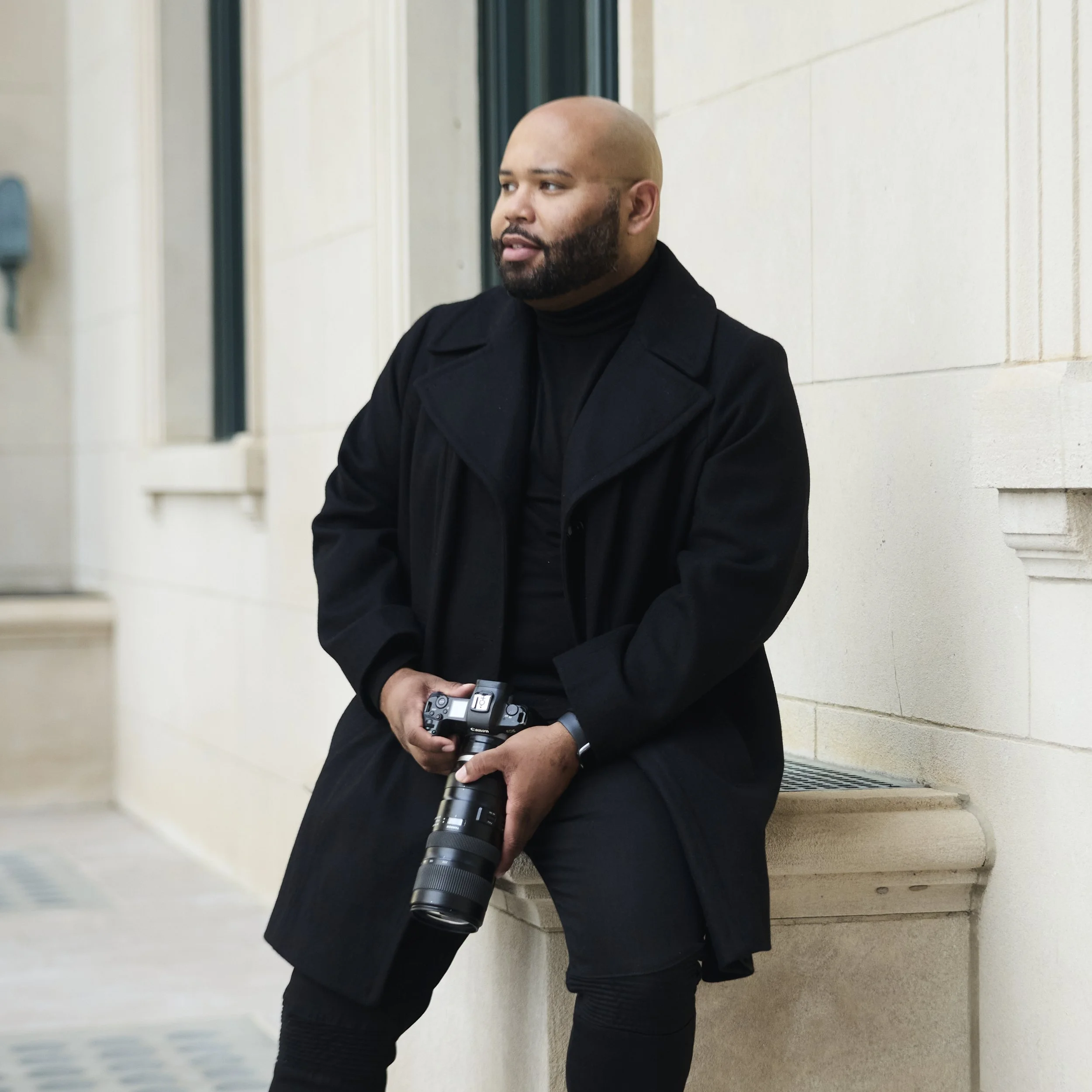 A man with a shaved head and beard wearing a black coat holding a camera, standing against a beige stone wall.