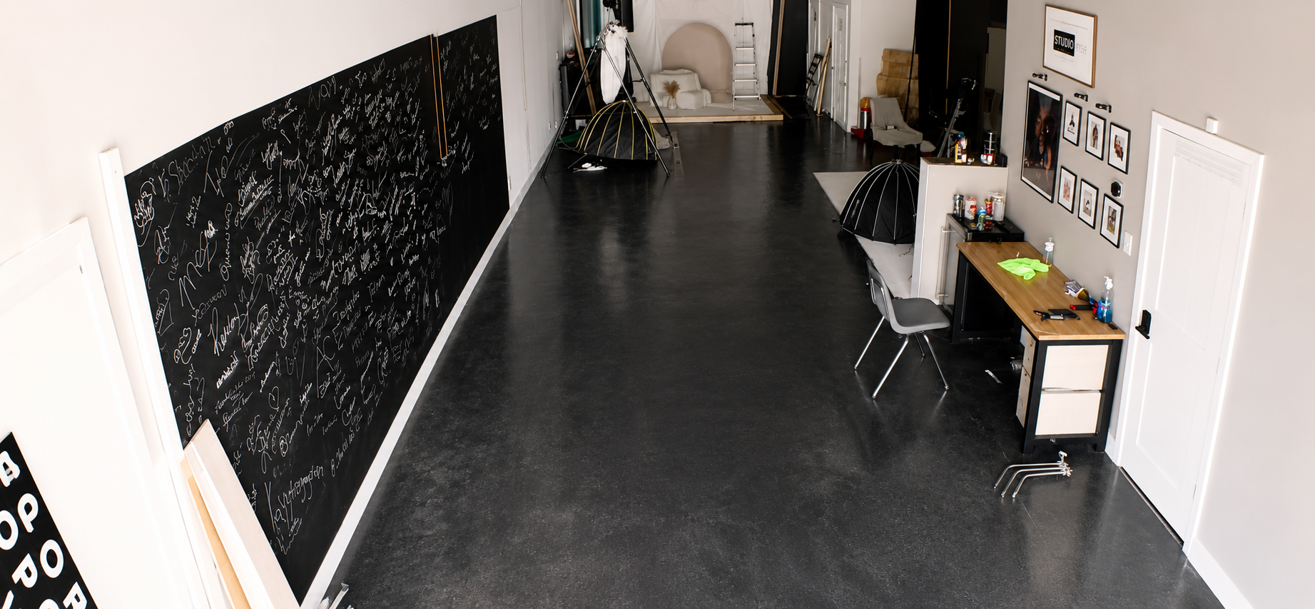 Interior of a modern art studio or workspace with a large blackboard wall filled with handwritten notes on the left, and a workspace with chairs, tables, and framed artwork on the right, featuring a black and white color scheme.