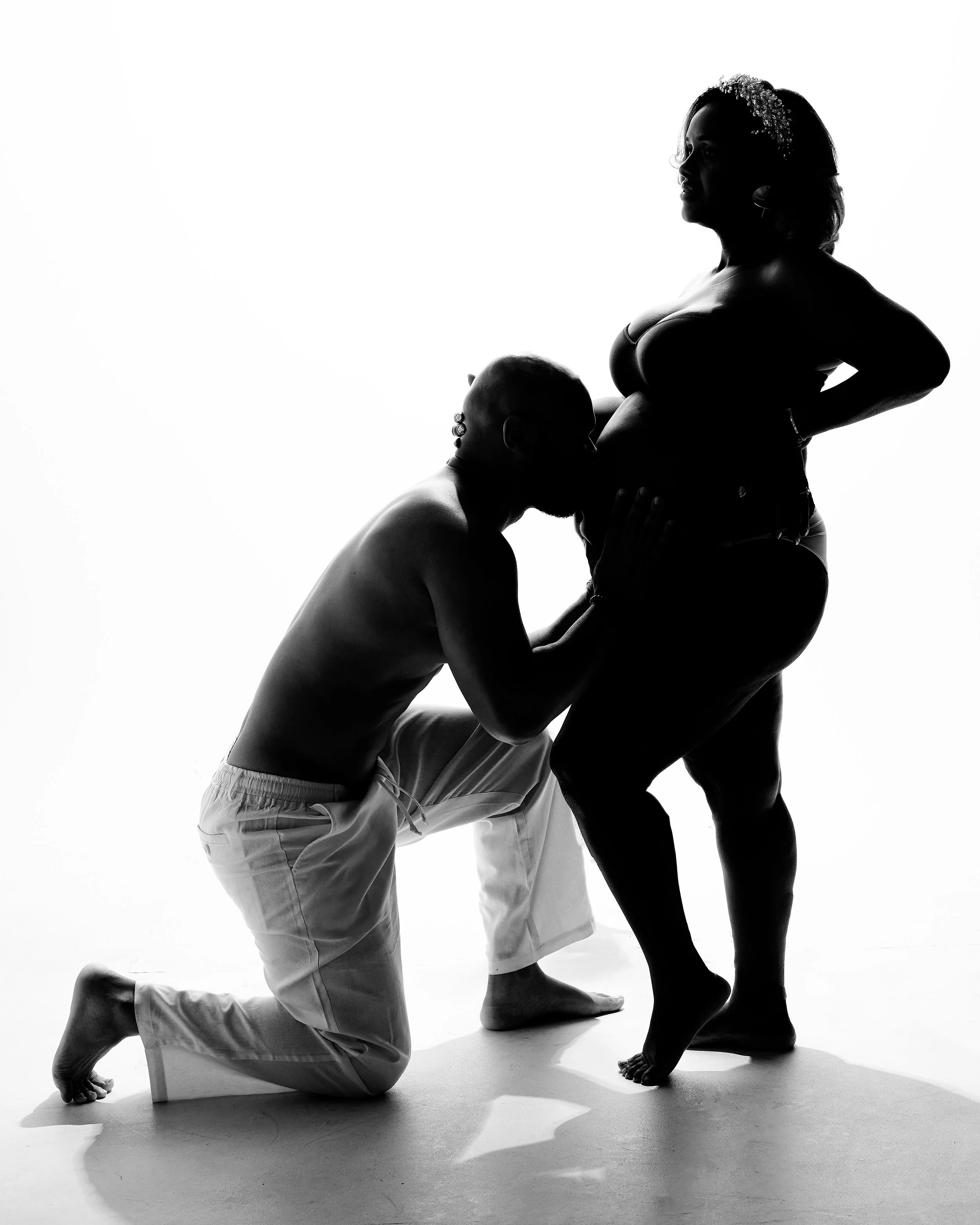 A black and white silhouette of a woman standing with her hands on her hips and a man kneeling with his head pressed against her abdomen, both with shaved heads. The woman appears to have tattoos on her arm and a flower headpiece, and the man is shir