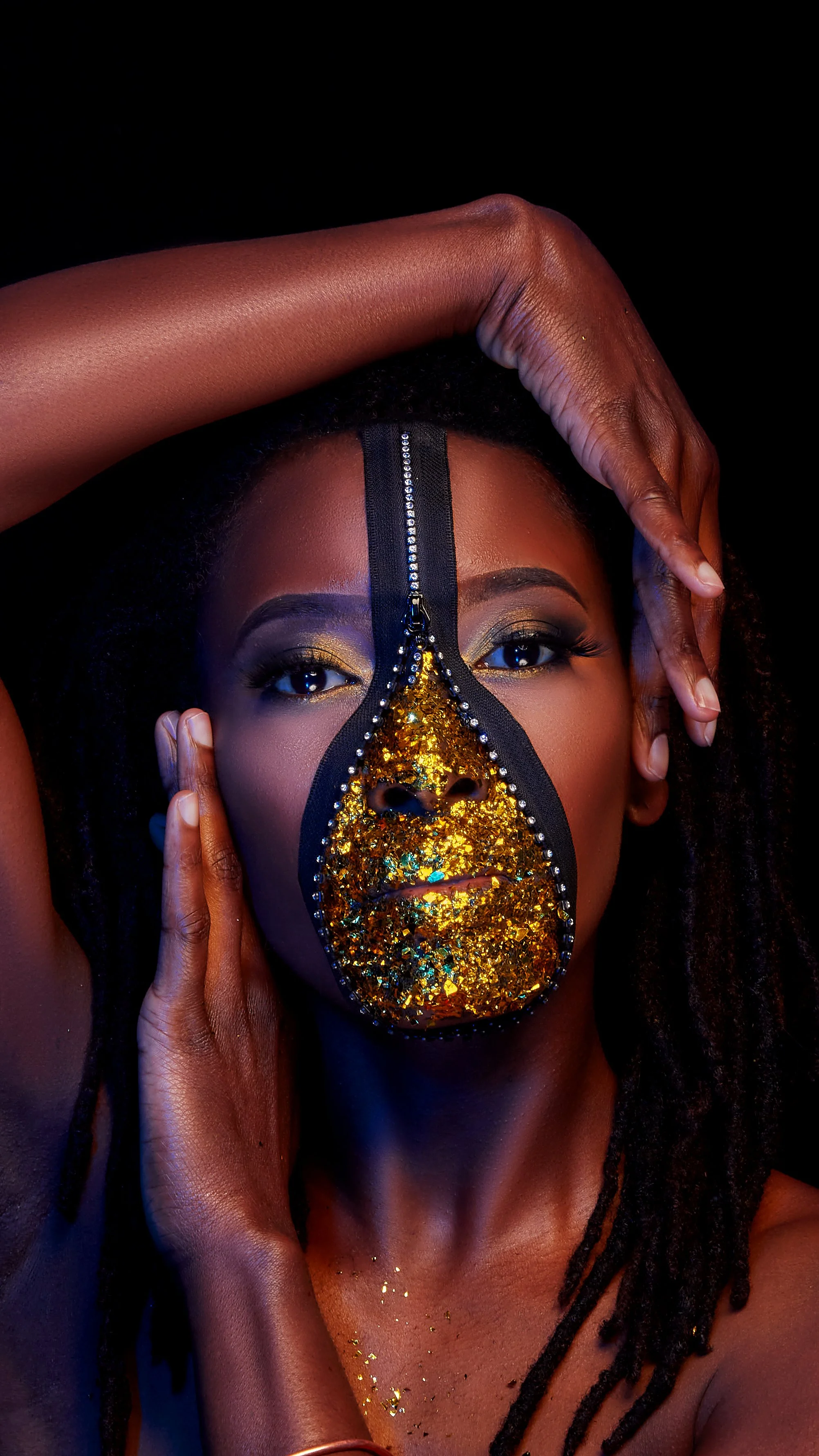 A woman with dark skin and curly hair poses with her hands framing her face. She wears makeup with gold eyeshadow and has a gold glittery mask covering her nose and lips, with a black border and silver rhinestones.