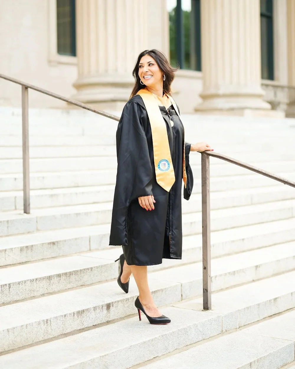 A woman in a black graduation gown and cap standing outside on stairs, smiling, with a gold stole, black heels, and a formal background of classical columns and building.
