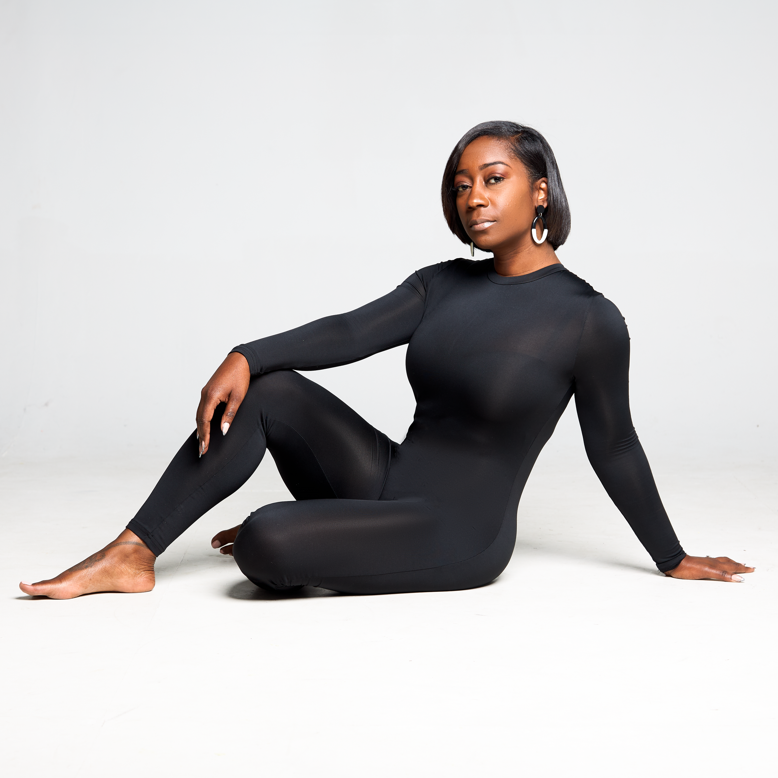 A woman with short black hair and dark skin is sitting on the floor against a white background. She is wearing a black long-sleeved top and black leggings. She has hoop earrings and is looking at the camera with a serious expression, her left arm res