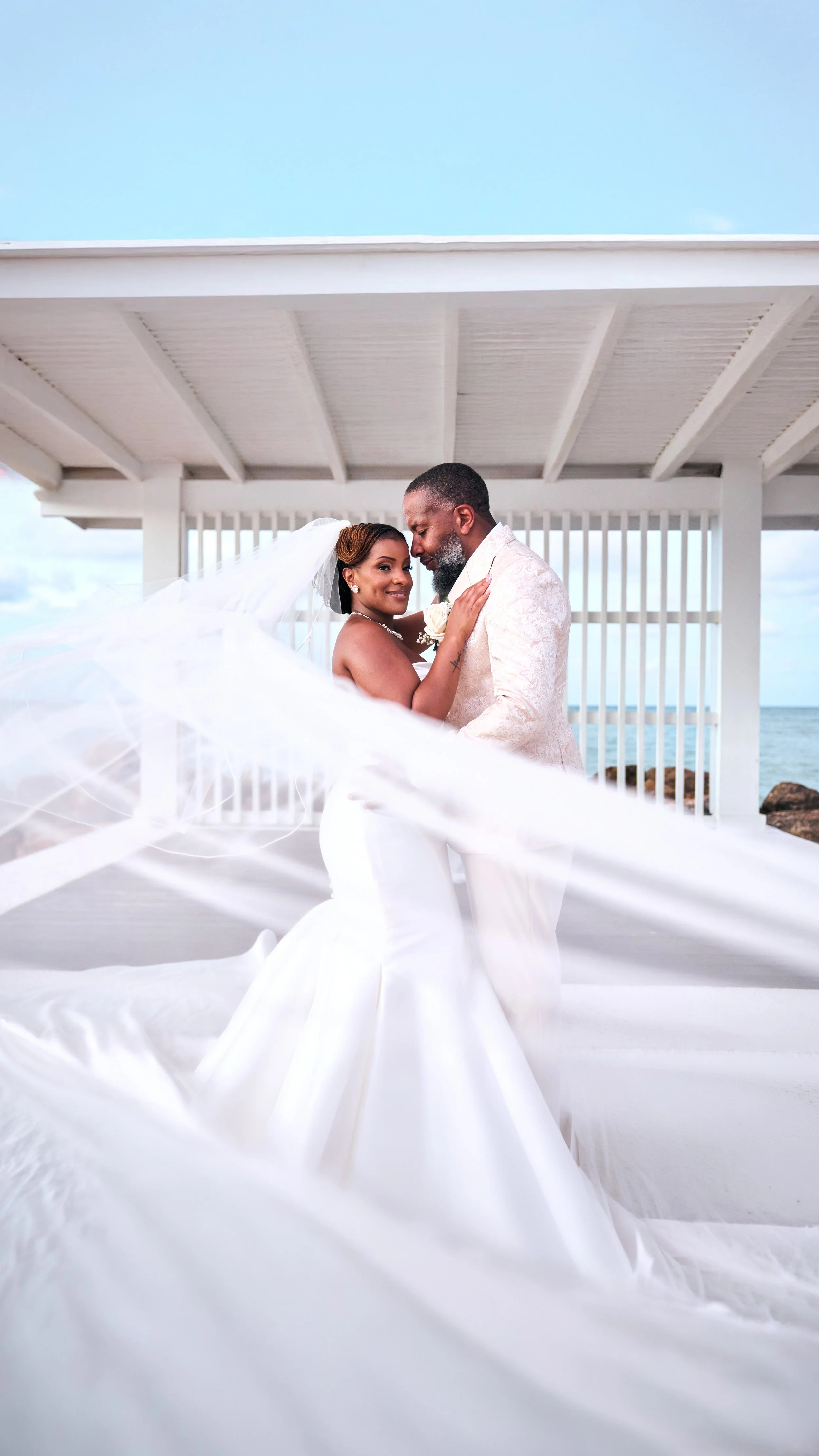 A bride and groom embracing on a white beachside pavilion, with the bride's flowing veil in the foreground and the ocean in the background.