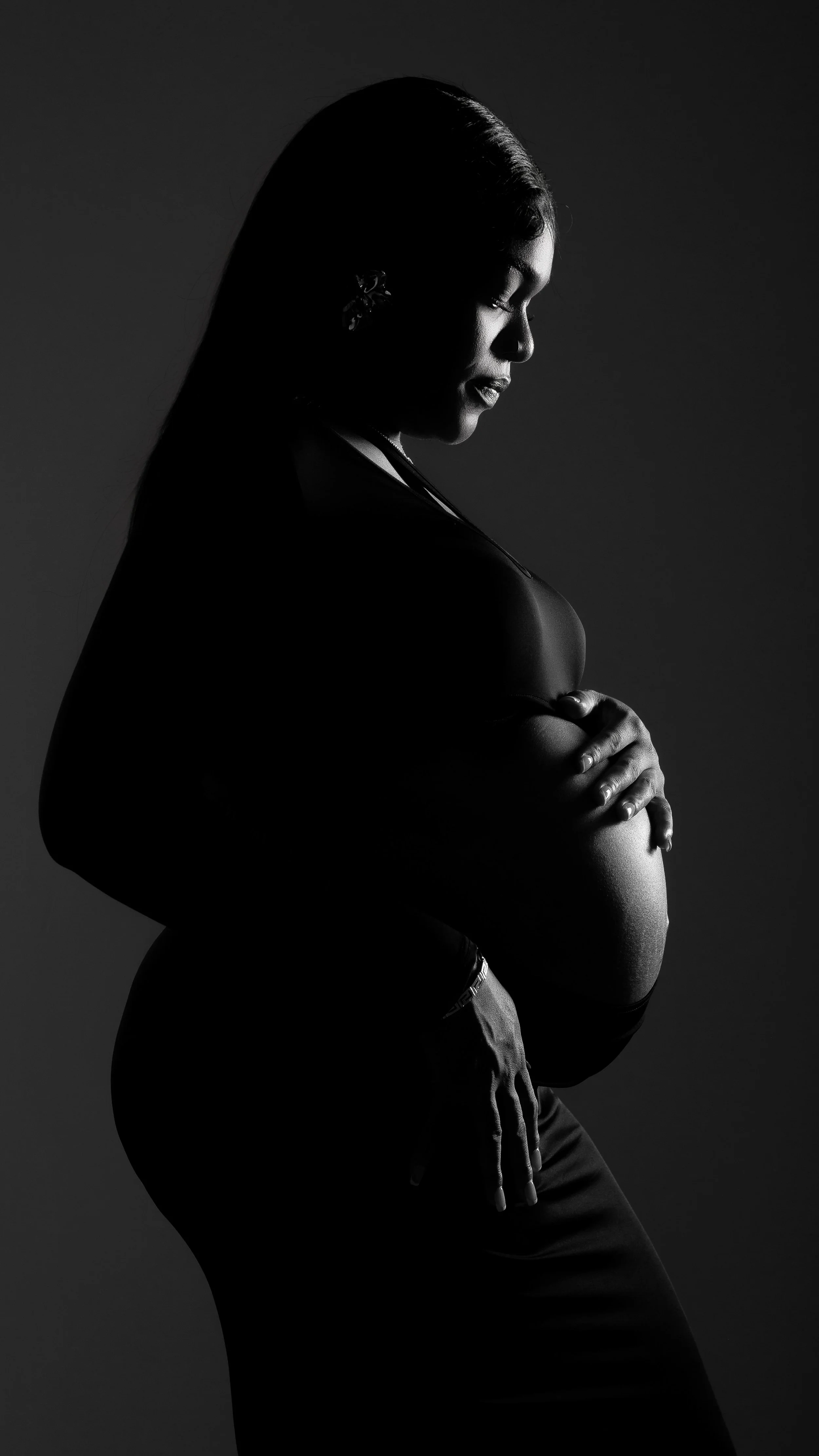 Black and white silhouette of a woman with her eyes closed, hugging her pregnant belly against a plain background.