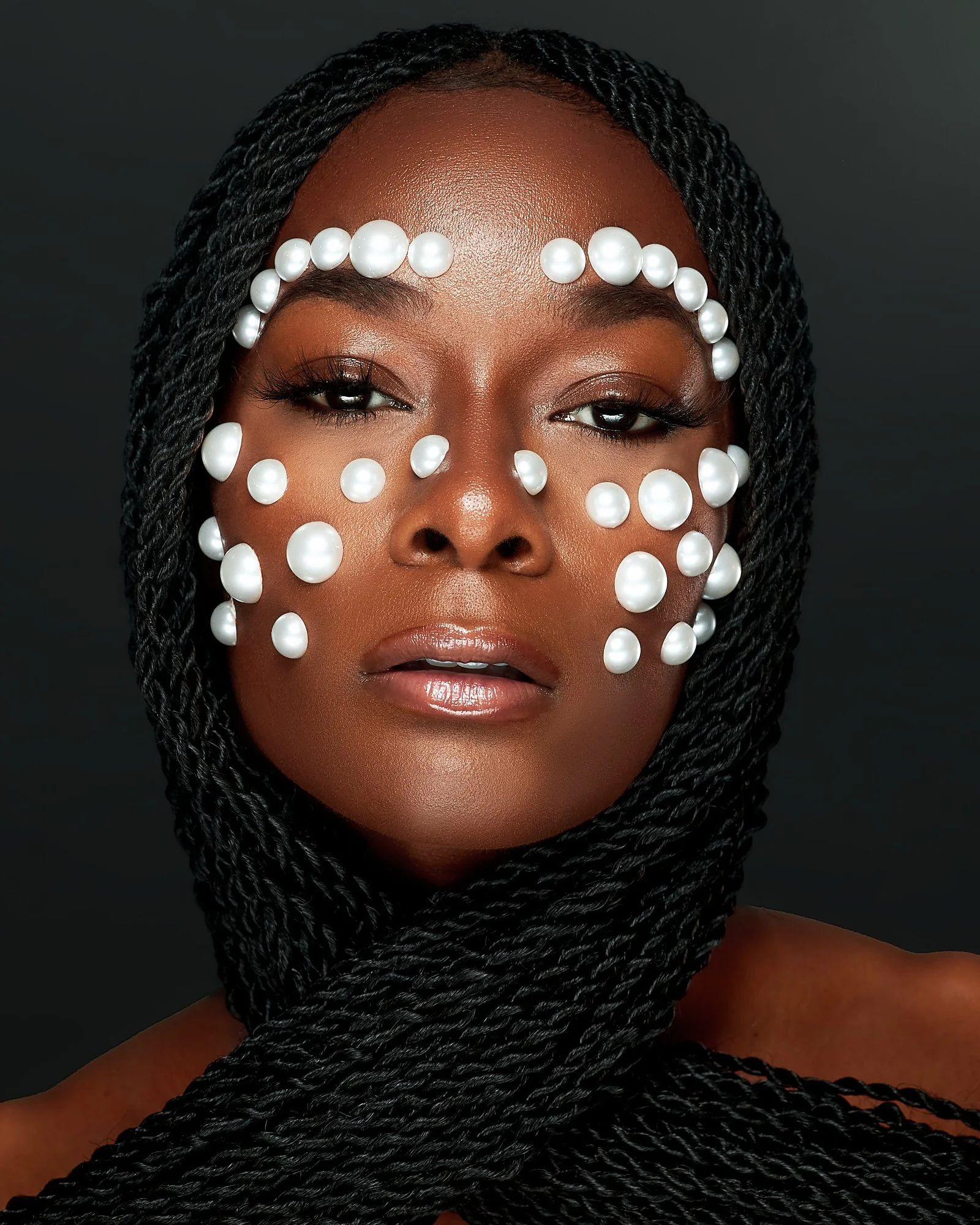 A woman with dark skin and braided hair, decorated with white pearls on her face and forehead, against a plain dark background.