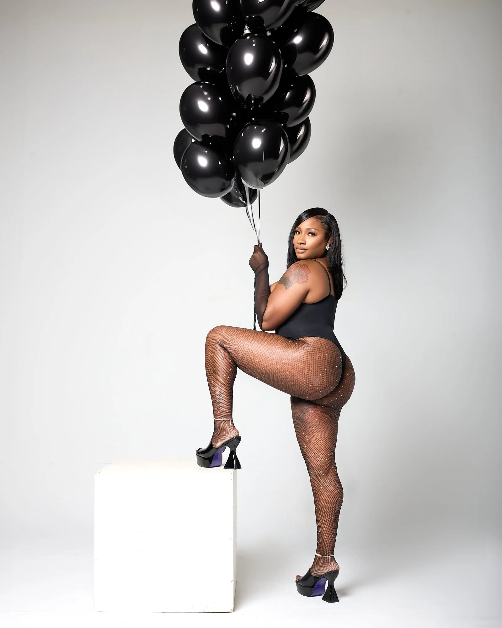 A woman with black hair and a tattoo on her shoulder poses with one leg raised on a small white platform. She wears a black outfit, fishnet stockings, and high platform shoes. She holds a bunch of black balloons and stands against a plain light backg