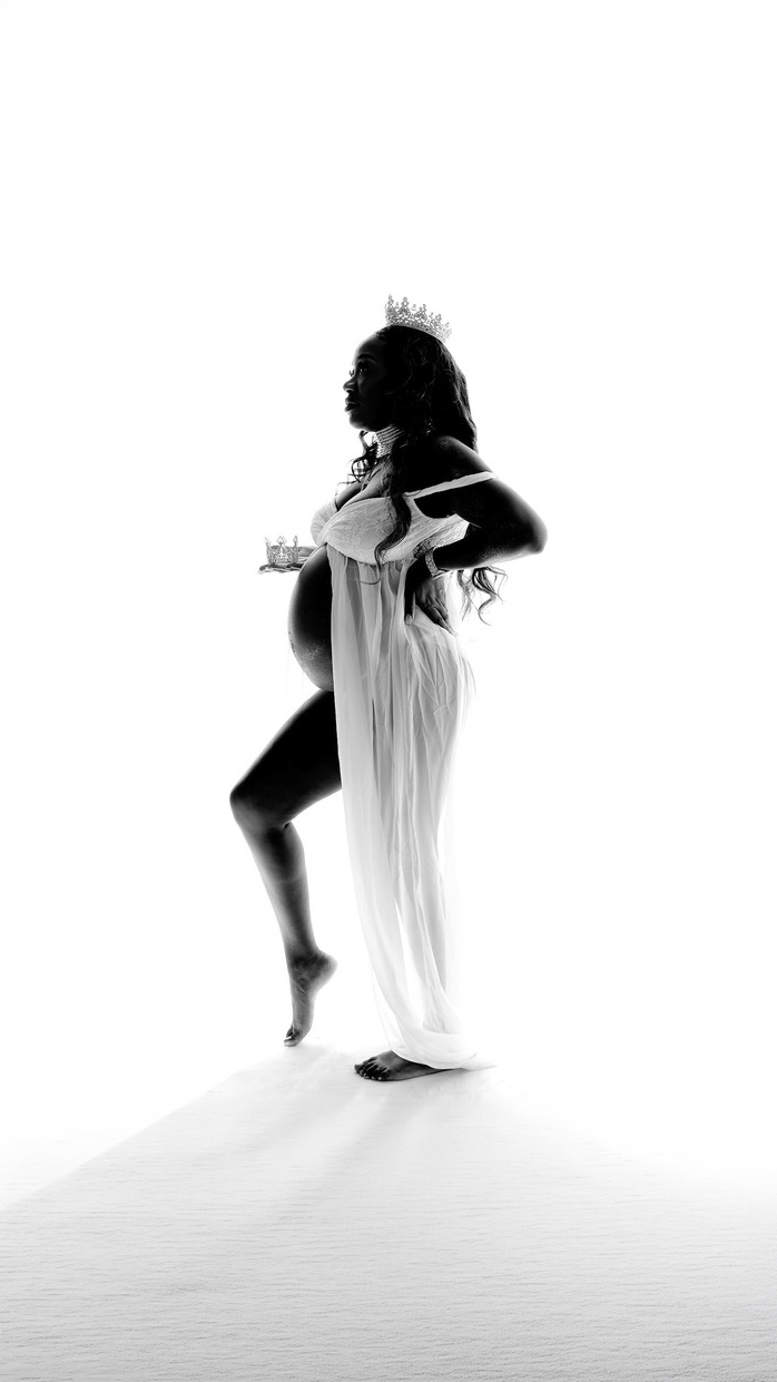 Silhouette of a pregnant woman wearing a crown and flowing dress, standing with one hand on her back and one foot slightly raised.