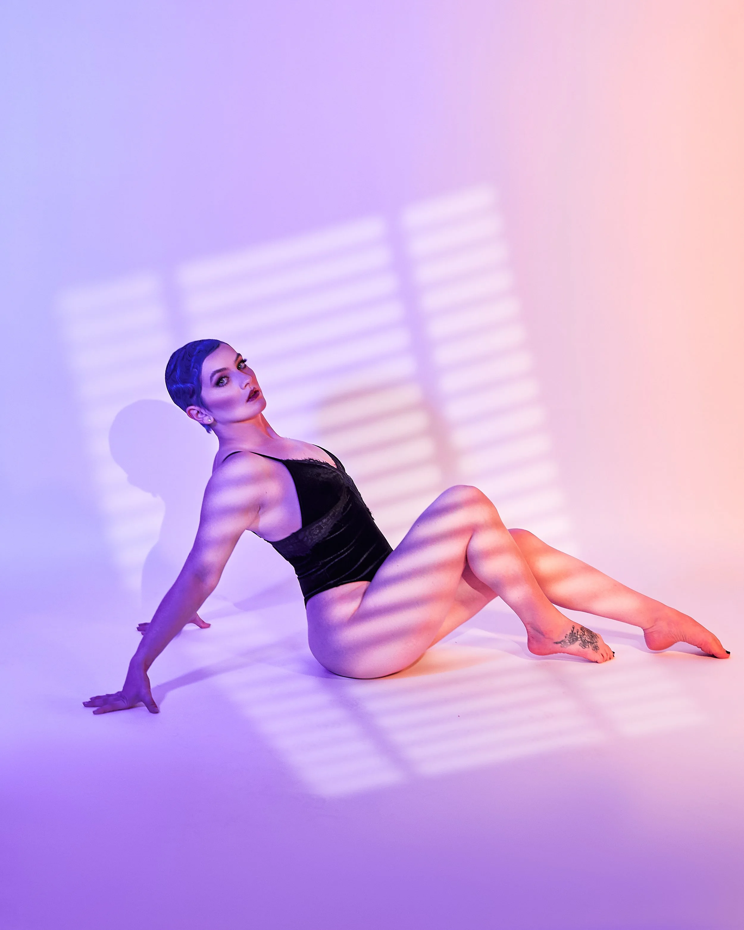 A woman with short dark hair and makeup, sitting on a white surface with her legs bent, wearing a black lace dress. The background is illuminated with pastel purple and pink lights, with shadow patterns on the wall.