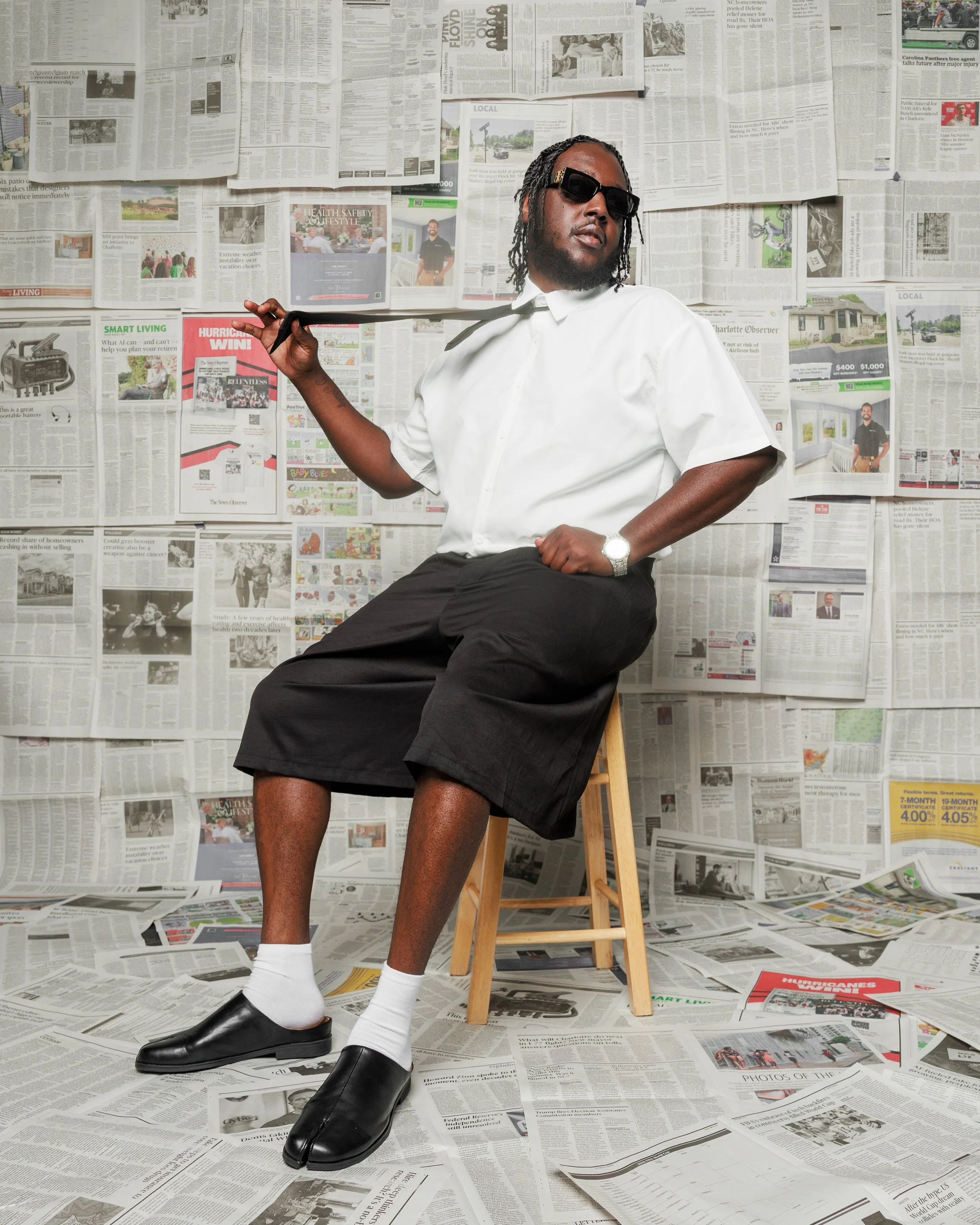 A man wearing sunglasses and a white shirt sits on a stool with his hand on his shoulder, posing in front of a backdrop of newspapers covered wall-to-wall, with newspapers scattered on the floor.