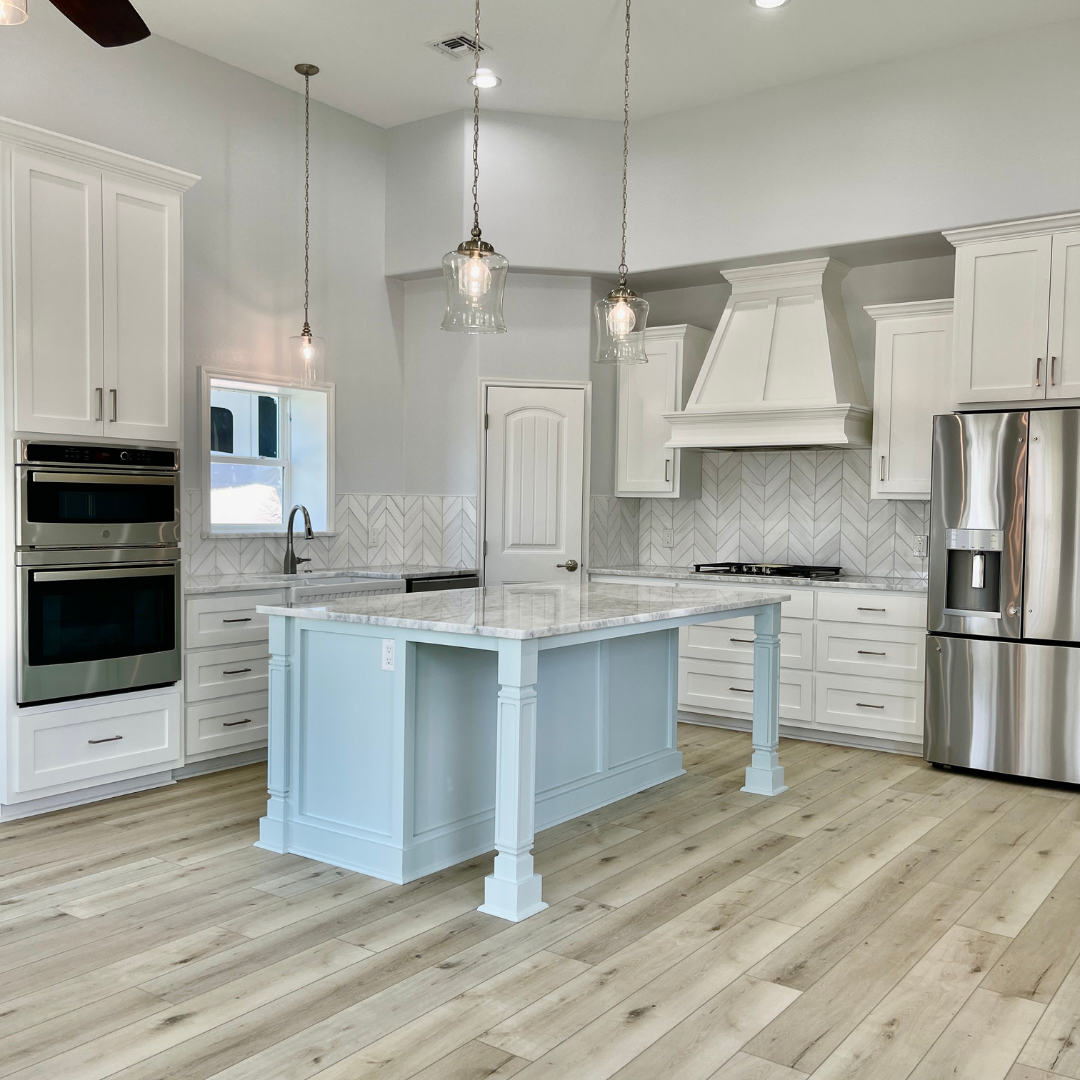 Modern white kitchen with stainless steel appliances, a large island with a marble top, pendant lighting, and light wood flooring.