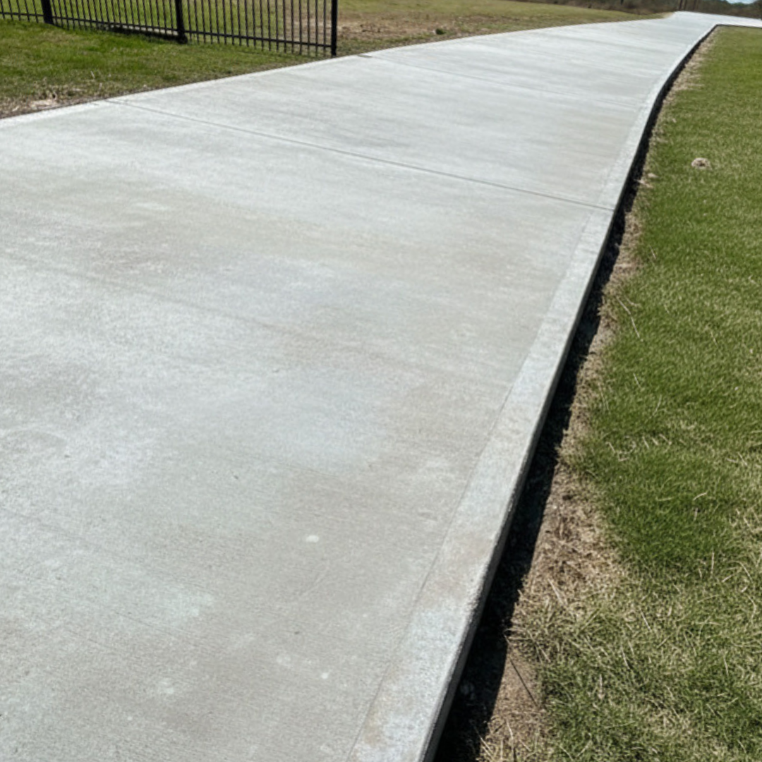 Concrete sidewalk with a slight incline, bordered by grass and a black metal fence.