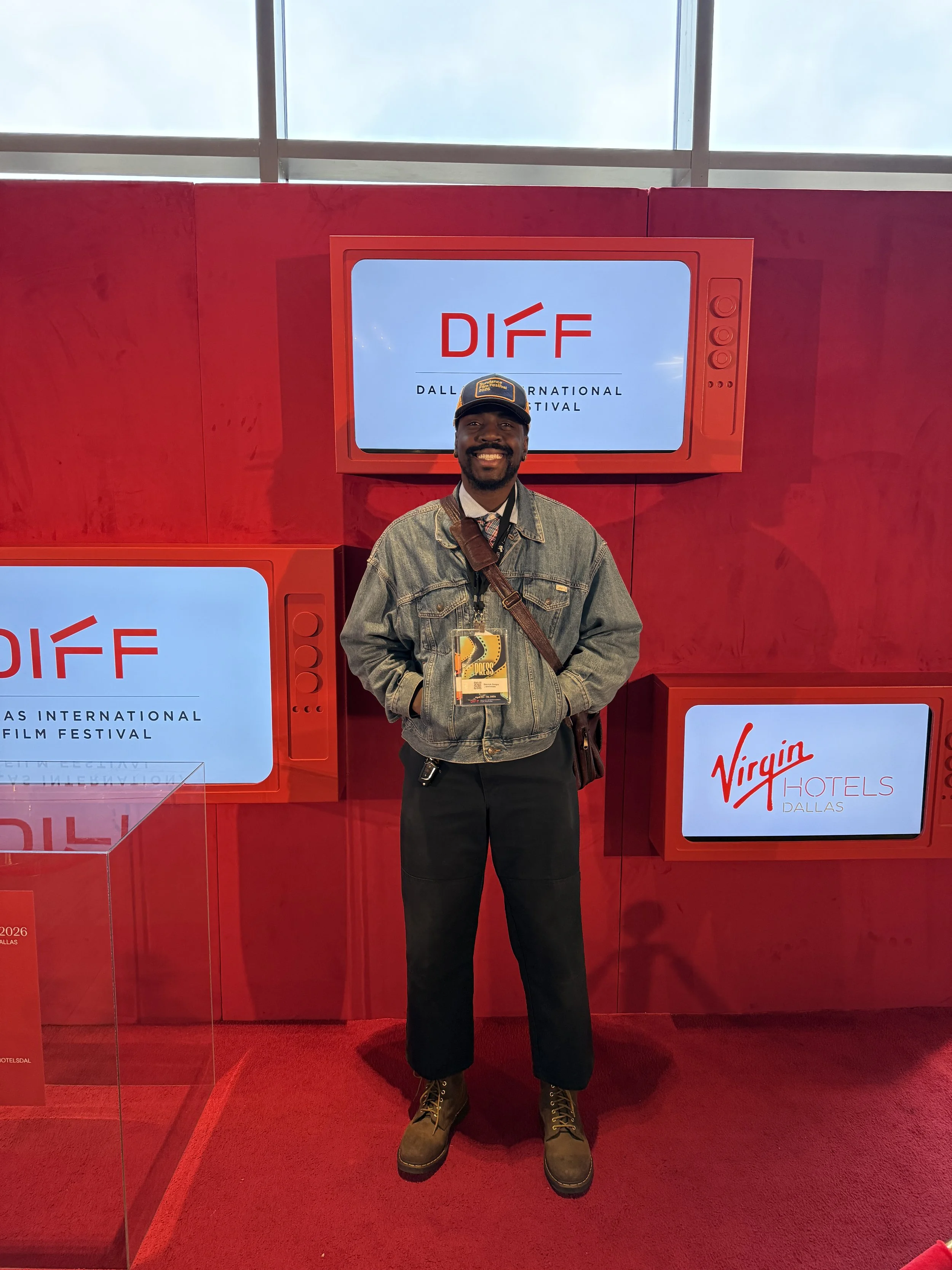 A man standing in front of a red wall with digital screens at the Dallas International Film Festival. He is smiling, wearing a denim jacket, black pants, brown boots, a cap, and has an event badge around his neck.