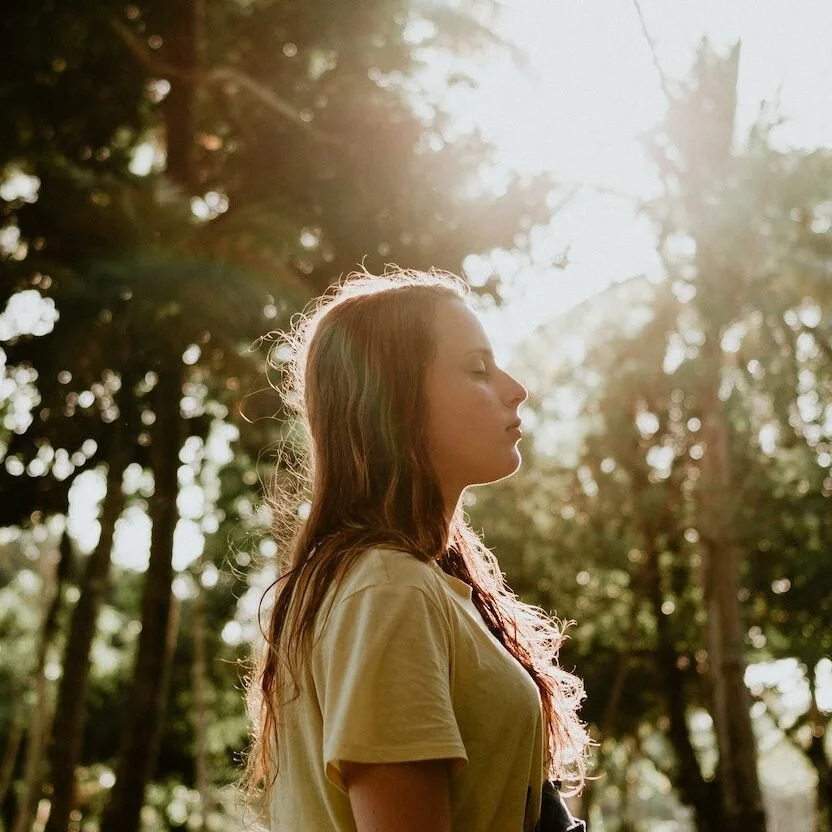 Woman standing in a sunlit forest with her eyes closed, reflecting calm and nervous system regulation.