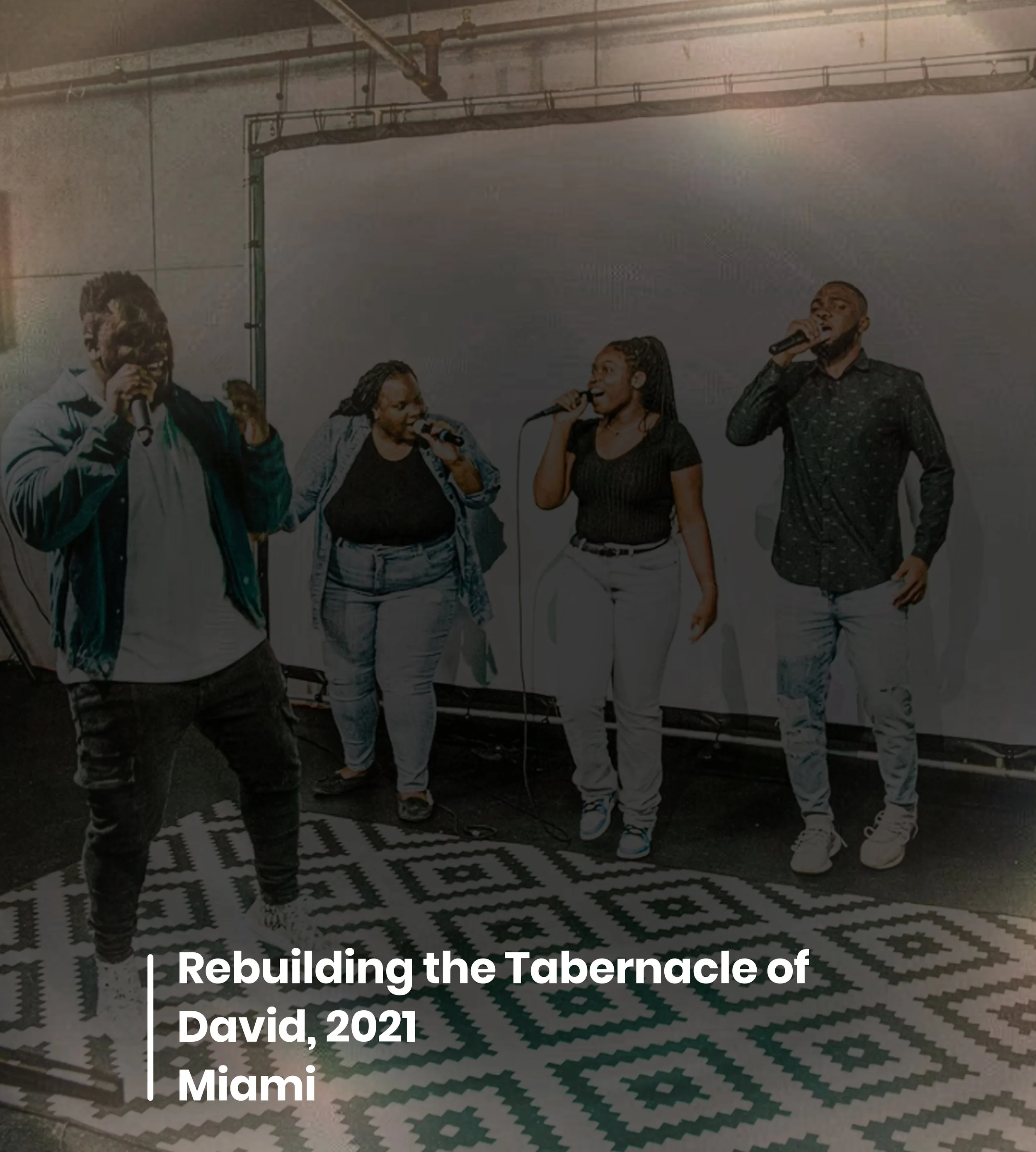 Group of five people singing into microphones on stage during a faith event, with a patterned floor and an industrial ceiling in the background.