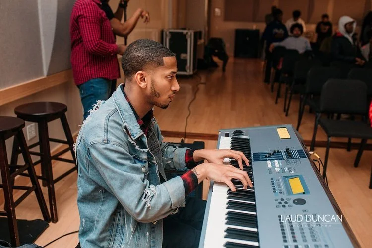 A young man playing a keyboard in a music rehearsal room with other people sitting and standing in the background.