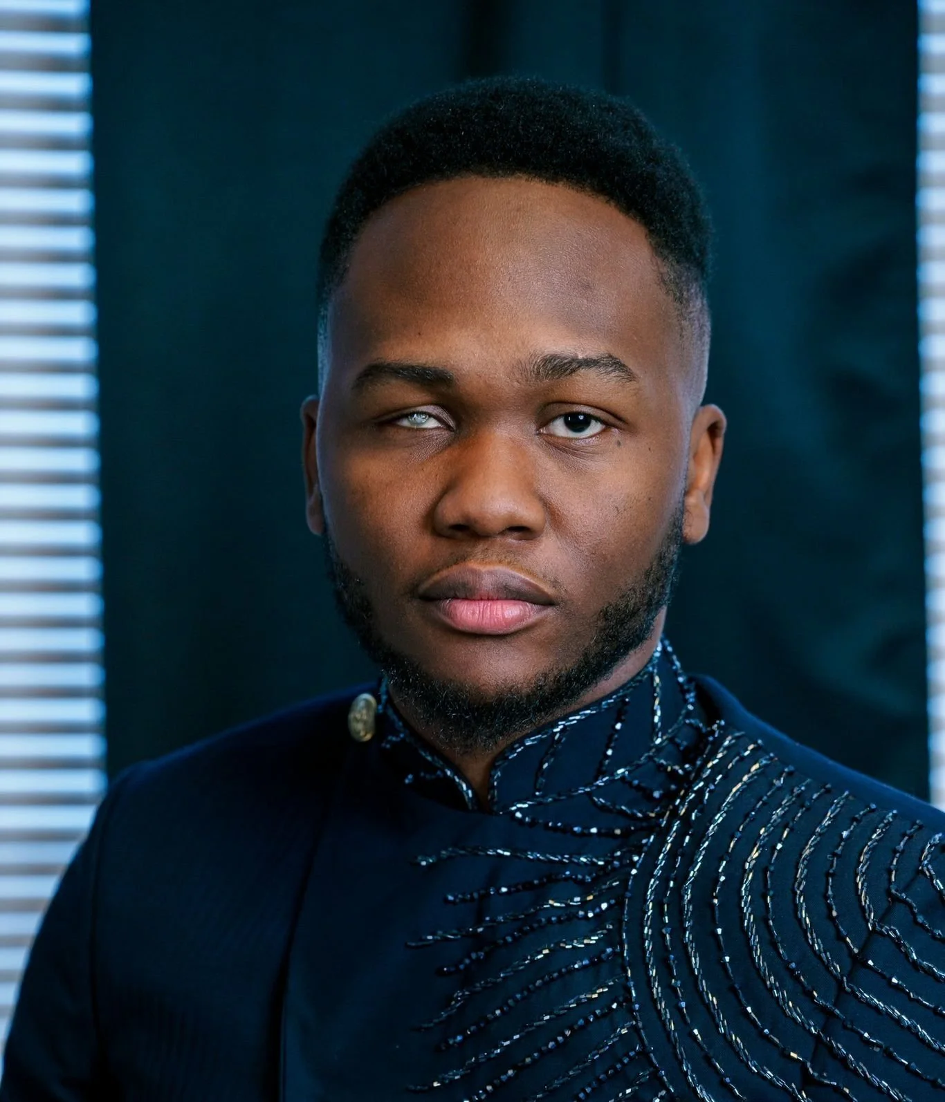 A young man with dark skin and short curly hair, wearing a dark, patterned shirt with a high collar, looking directly at the camera with a serious expression, against a background of dark and light shades with vertical blinds.