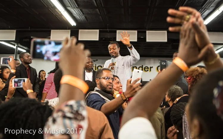 Man speaking into a microphone on stage while people in the audience take photos and clap.