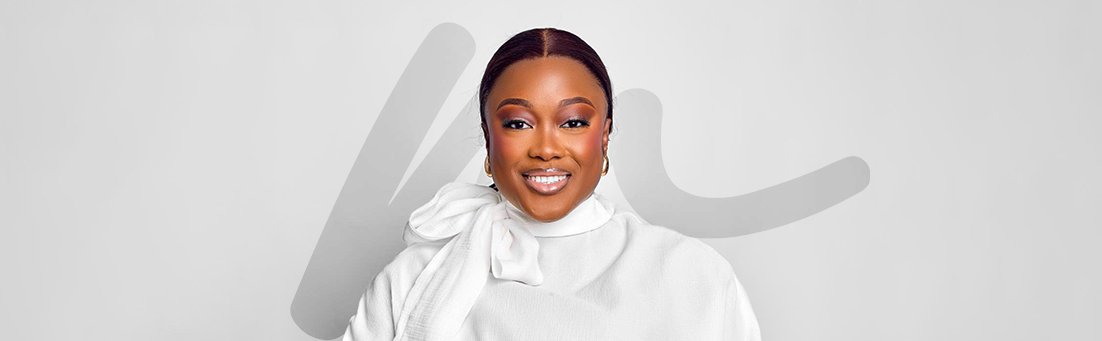 Smiling woman with dark hair wearing a white outfit with a large bow, posing in front of a light gray background.