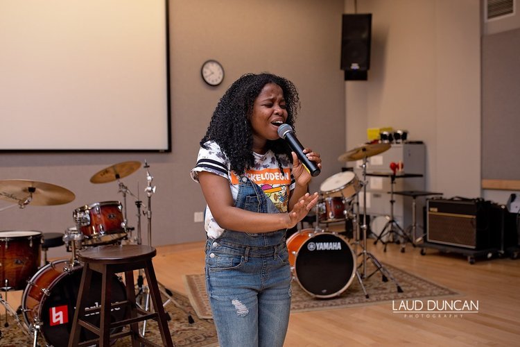 A young woman singing into a microphone in a music studio with drums in the background.