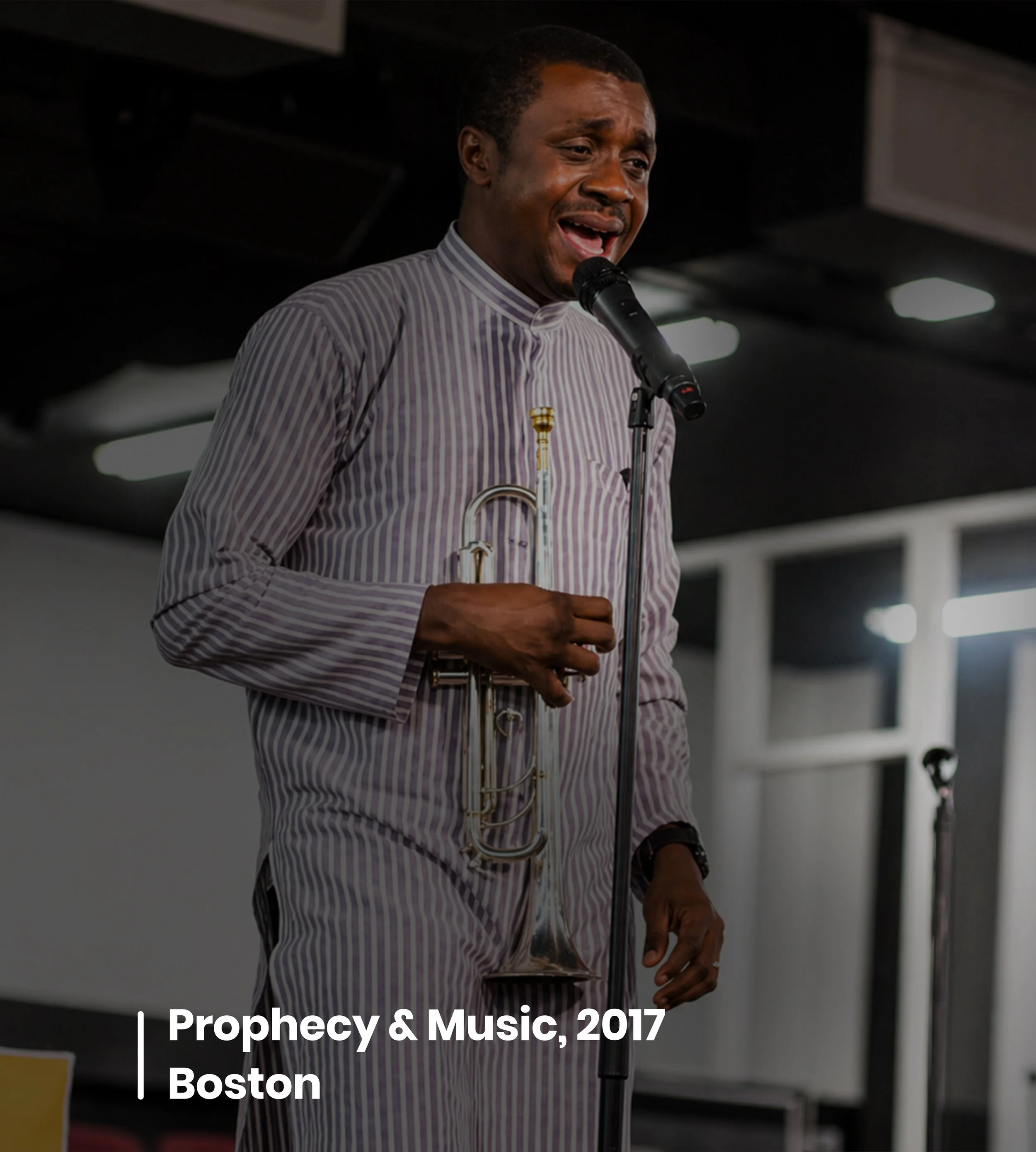 A man holding a microphone and a trumpet, speaking at an event titled 'Prophecy & Music, 2017' in Boston.