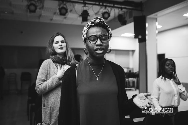 A woman with glasses and a headwrap appears to be singing or praying with eyes closed, with two other women in the background, in an indoor setting.