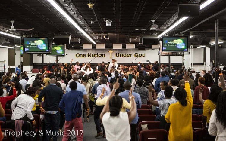 Crowd of people in a church holding their hands up during worship service, with a stage at the front featuring a large sign that reads 'One Nation Under God,' and screens displaying song lyrics.