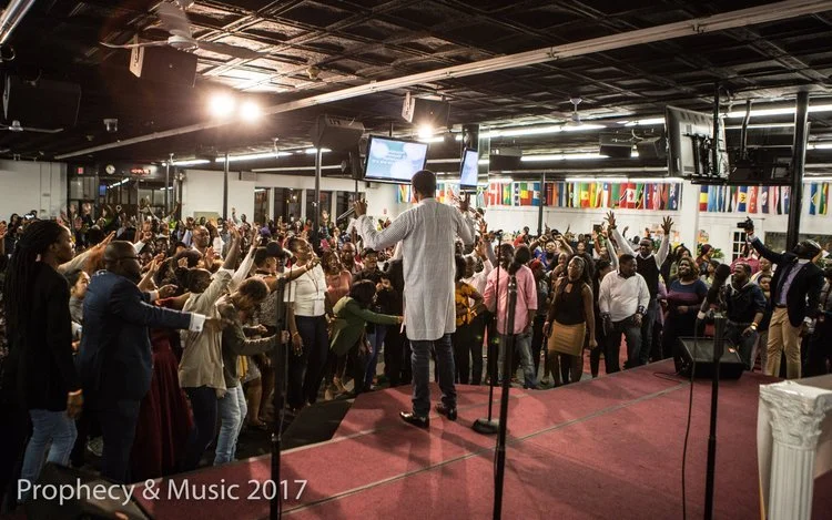 A large indoor religious gathering or church service with a preacher on stage leading the congregation in prayer or worship, many people with hands raised, flags from various countries hanging from the ceiling, and multiple screens display text or im