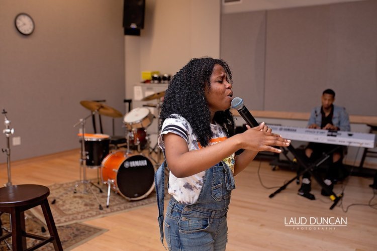A young girl with curly black hair singing into a microphone, with a man playing a keyboard in the background and a drum set nearby in a music studio or practice room.