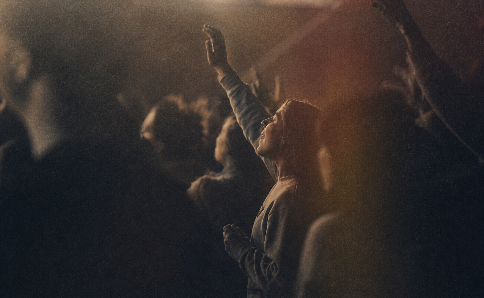 A woman at a concert with eyes closed and one hand raised, smiling as she enjoys the music, with other crowd members in the background.