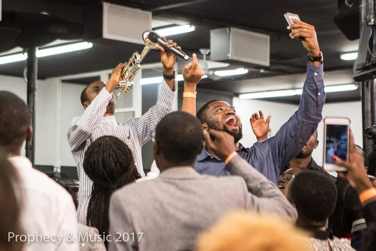A group of people at a lively event, with two men at the front taking selfies and one man holding a microphone, smiling and celebrating.