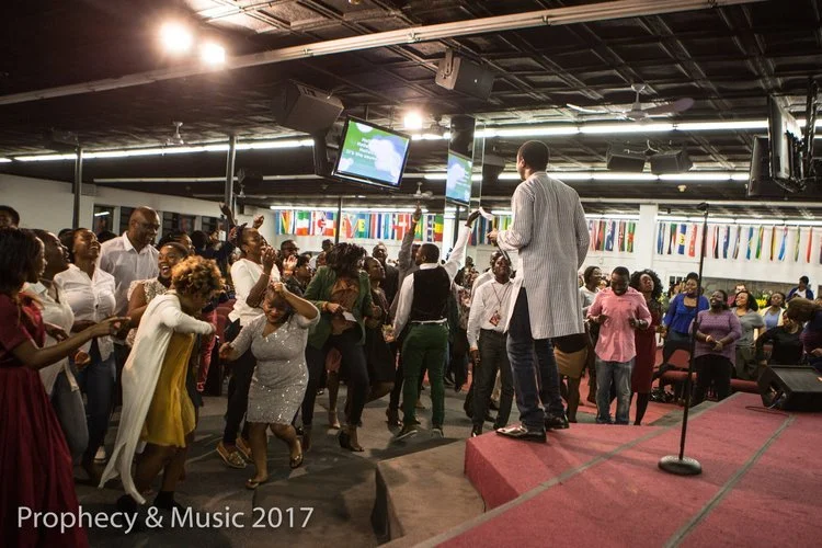 A diverse group of people dancing and enjoying a celebration in a crowded indoor venue with flags hanging in the background and a man speaking or singing on a stage.