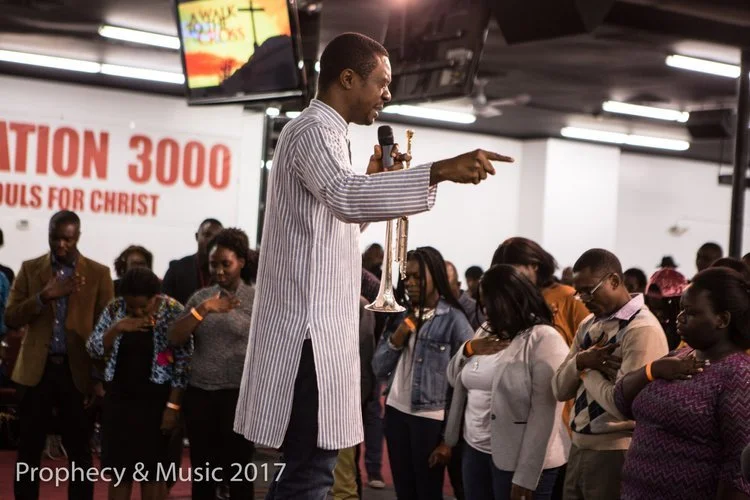 A preacher holding a microphone and pointing, standing on a stage in front of a congregation inside a church during a religious service.