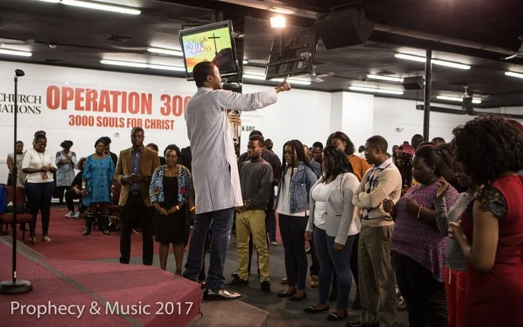 A man speaking to a group of people during a church event called Operation 300, with a banner reading 3000 Souls for Christ in the background.