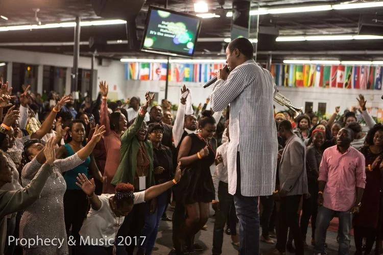 A man holding a microphone performs music to a crowd raising their hands and dancing in an indoor setting with international flags hanging from the ceiling.