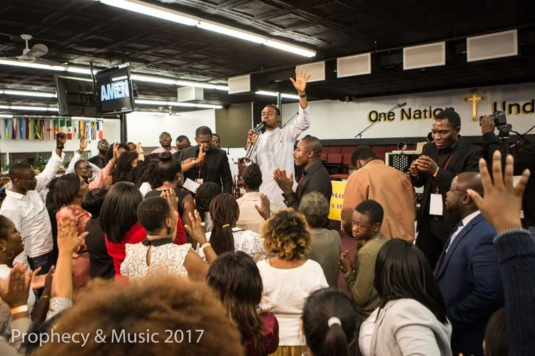 A man speaking passionately into a microphone at an indoor gathering, with a crowd raising hands and clapping in front of him. The background shows a sign that says 'One Nation Under God,' and American flags are hanging on the left side. The image is