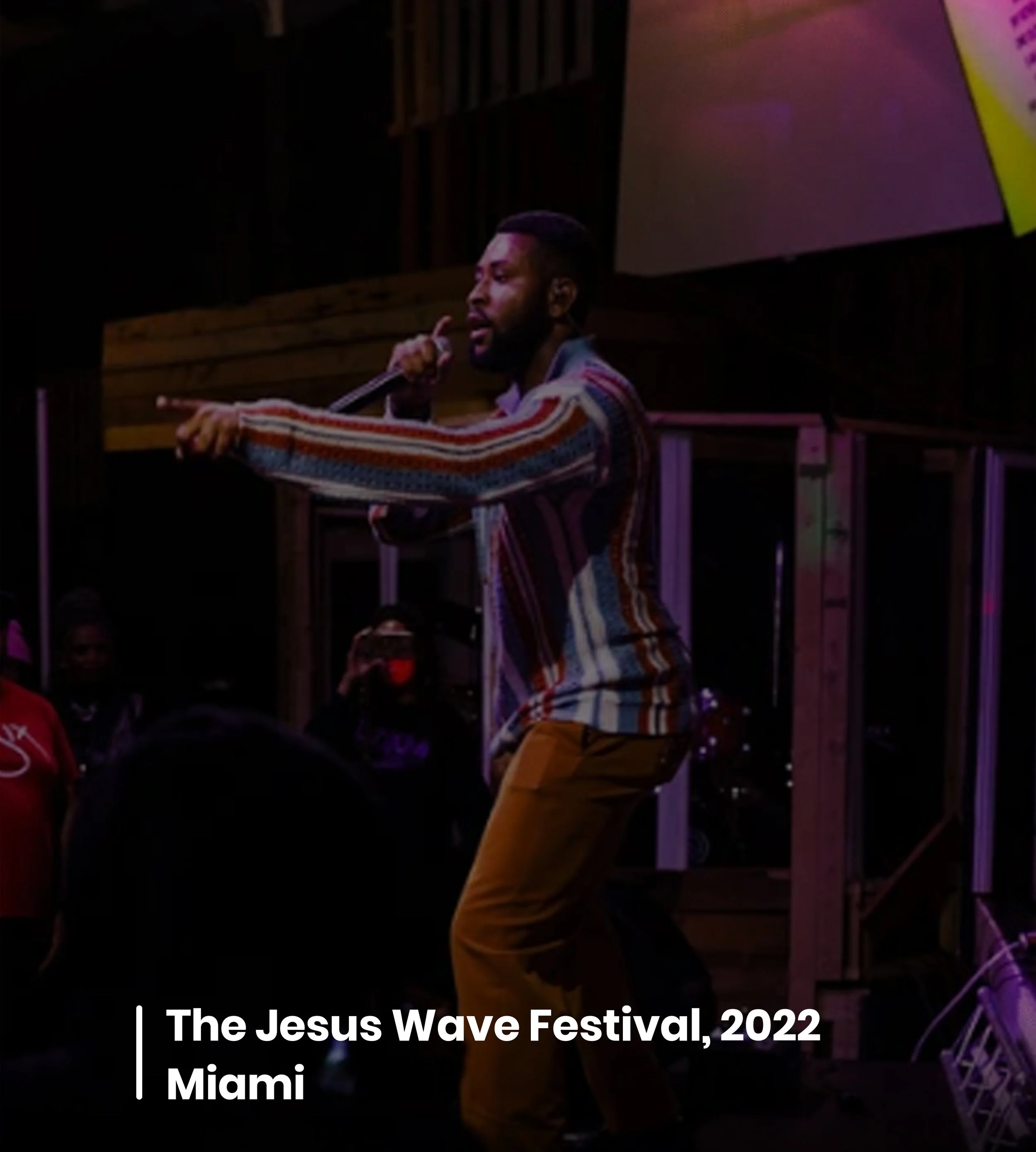 A man with a beard and mustache, wearing a colorful striped long-sleeve shirt and mustard-colored pants, is speaking or singing into a microphone on stage at an event called The Jesus Wave Festival 2022 in Miami. The background features a large screen with colorful visuals and some audience members are visible in the foreground.