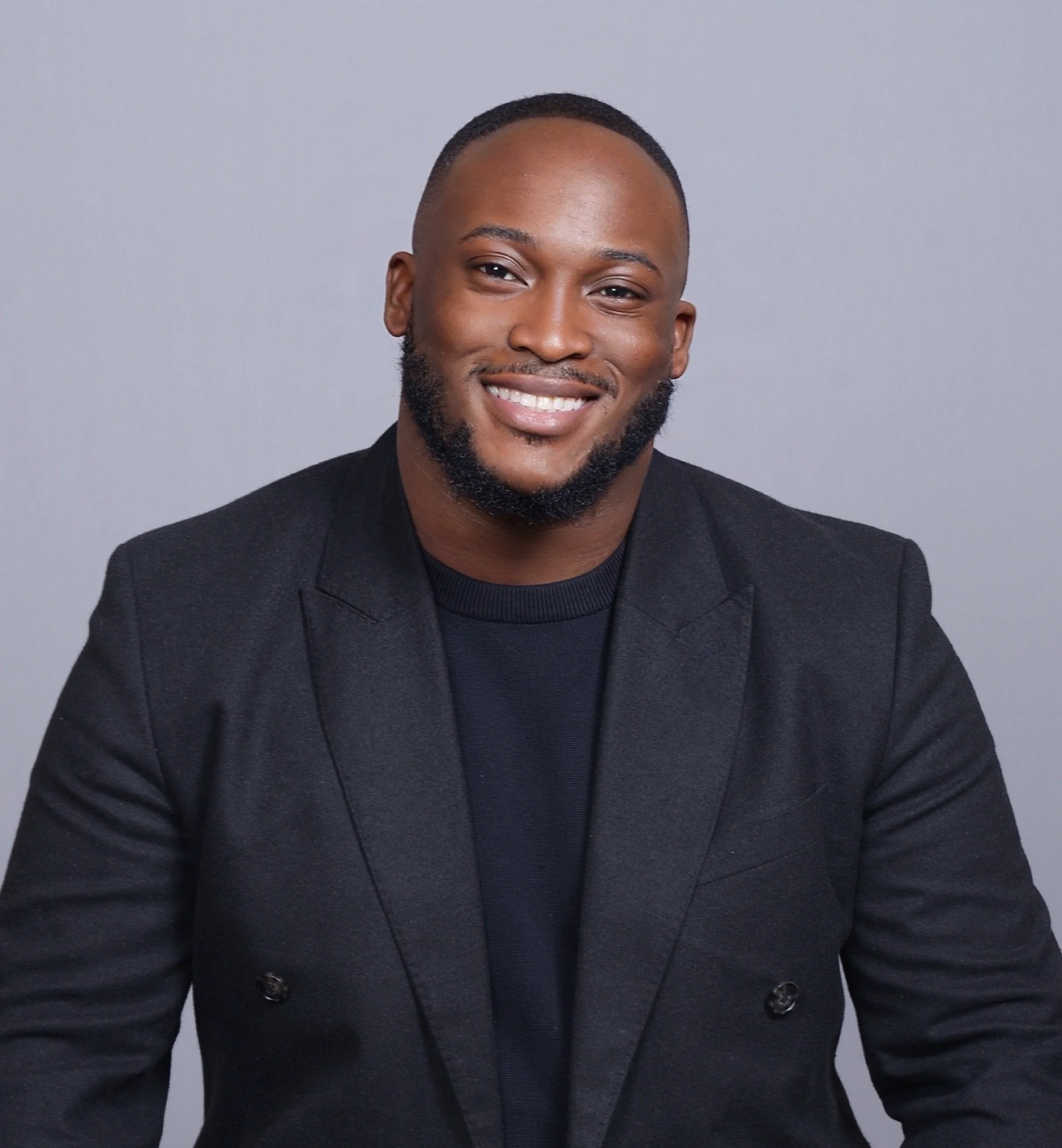 Close-up of a smiling man with a short haircut, beard, and dark skin, wearing a black blazer and shirt, against a plain gray background.