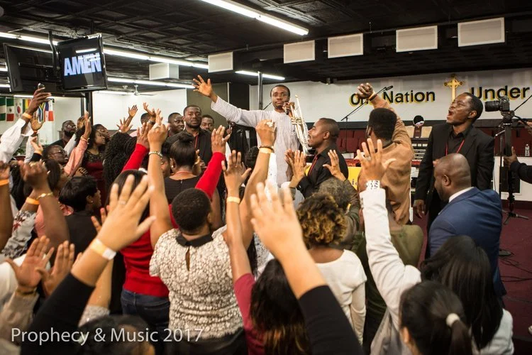 A crowd of people gathered in a church setting with a man leading worship or prayer. Many individuals have their hands raised, and the man at the front appears to be speaking or praying with him. The room has church banners and a screen displaying 'A
