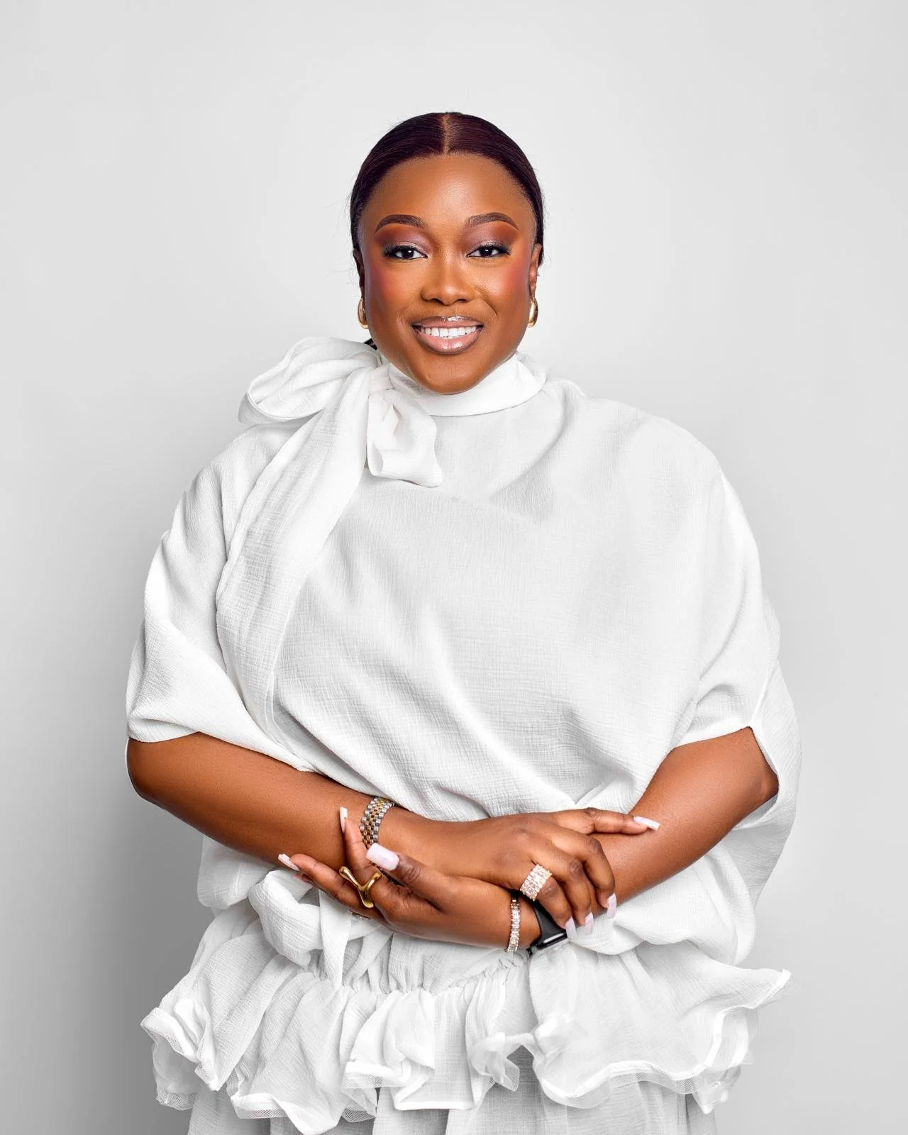 A woman smiling in a white dress with ruffled details and a bow on the shoulder, standing against a gray background.