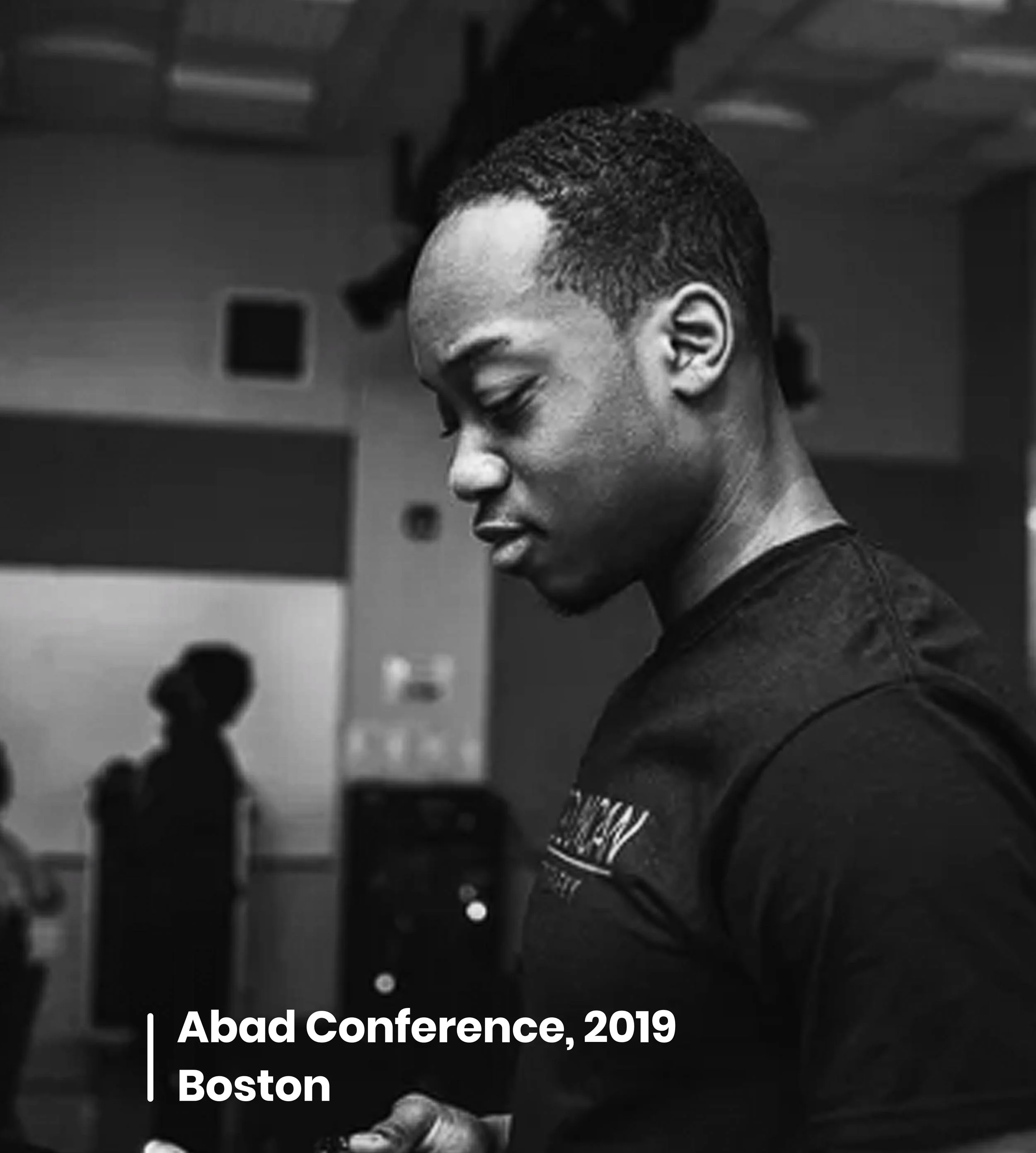 Black and white photo of a young man with short hair, looking down, with a conference banner in the background that reads 'Abad Conference, 2019 Boston'.