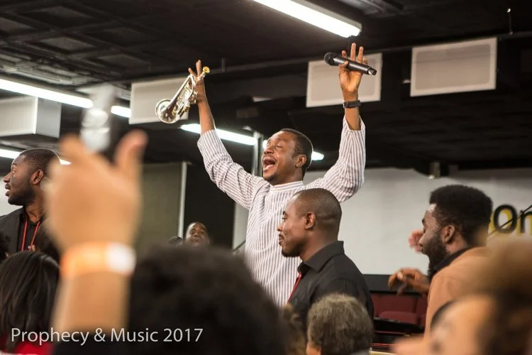 A man holding a microphone in one hand and a clarinet in the other, raising both in the air, appears to be singing or speaking passionately at an indoor event with a crowd.