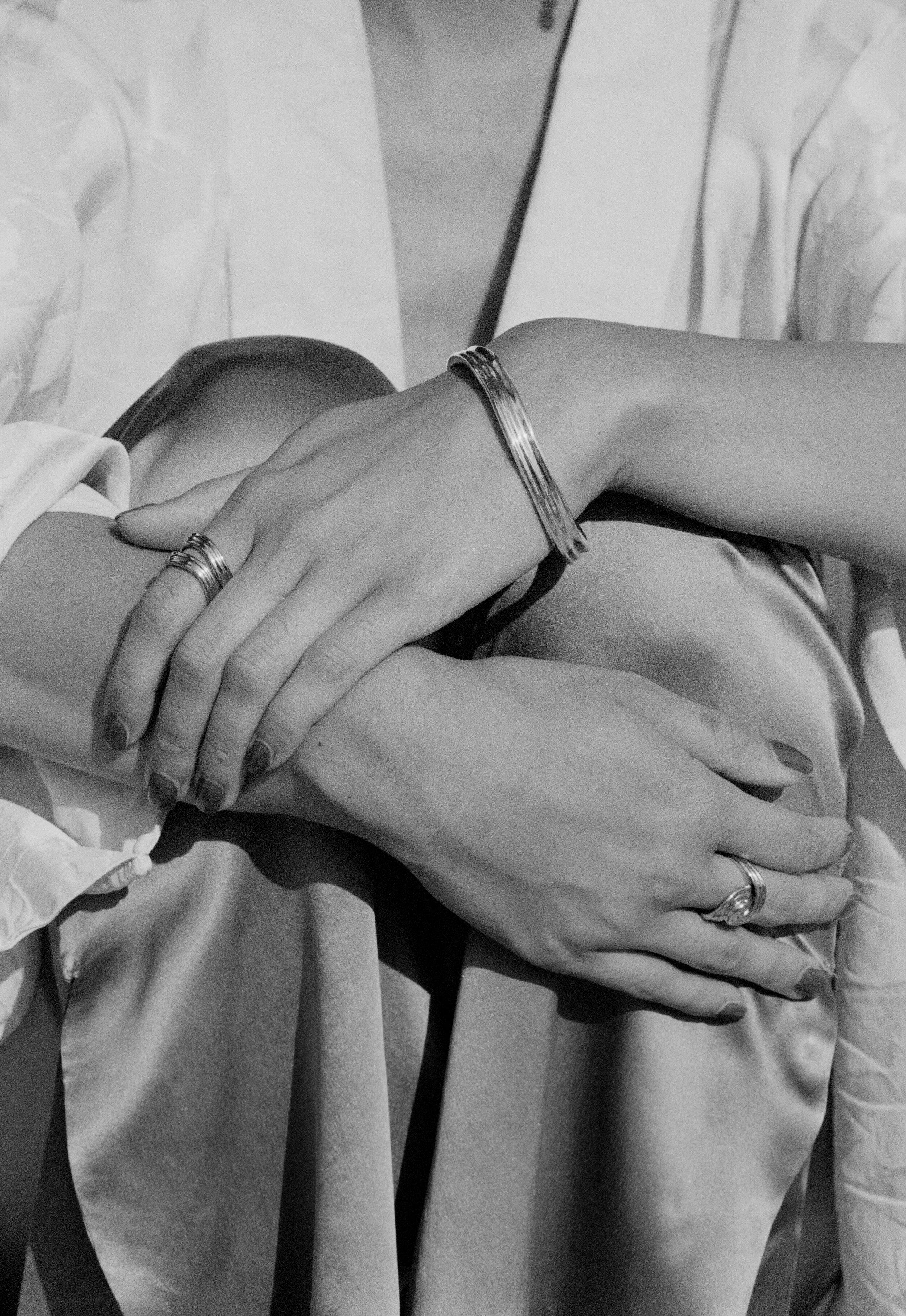 Two hands with rings and bracelets resting on a person's lap, black and white photograph.