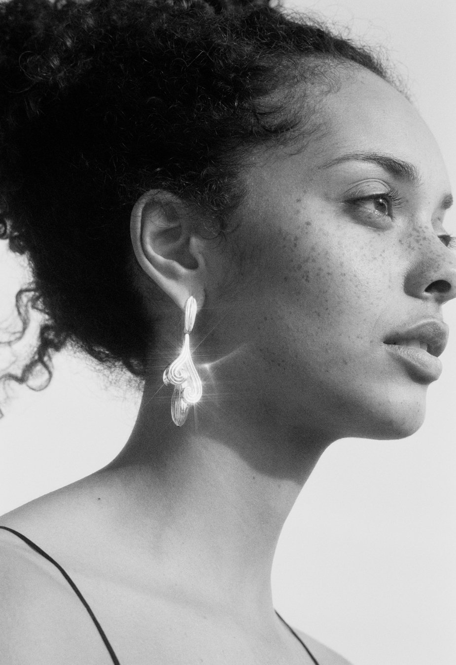 Black and white photo of a woman with curly hair, wearing a decorative earring, looking to the right.