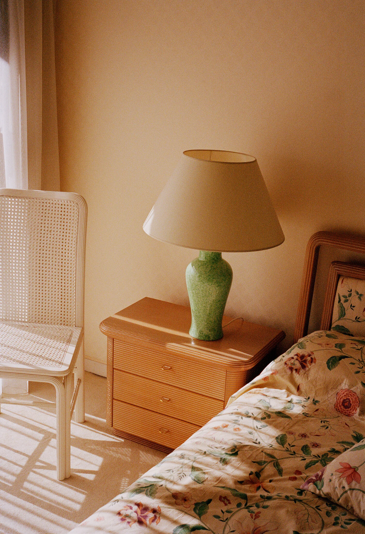 A bedroom scene with a floral bedspread, a wooden nightstand with a green ceramic table lamp, and a wicker chair near a window with sheer curtains, all bathed in warm sunlight.