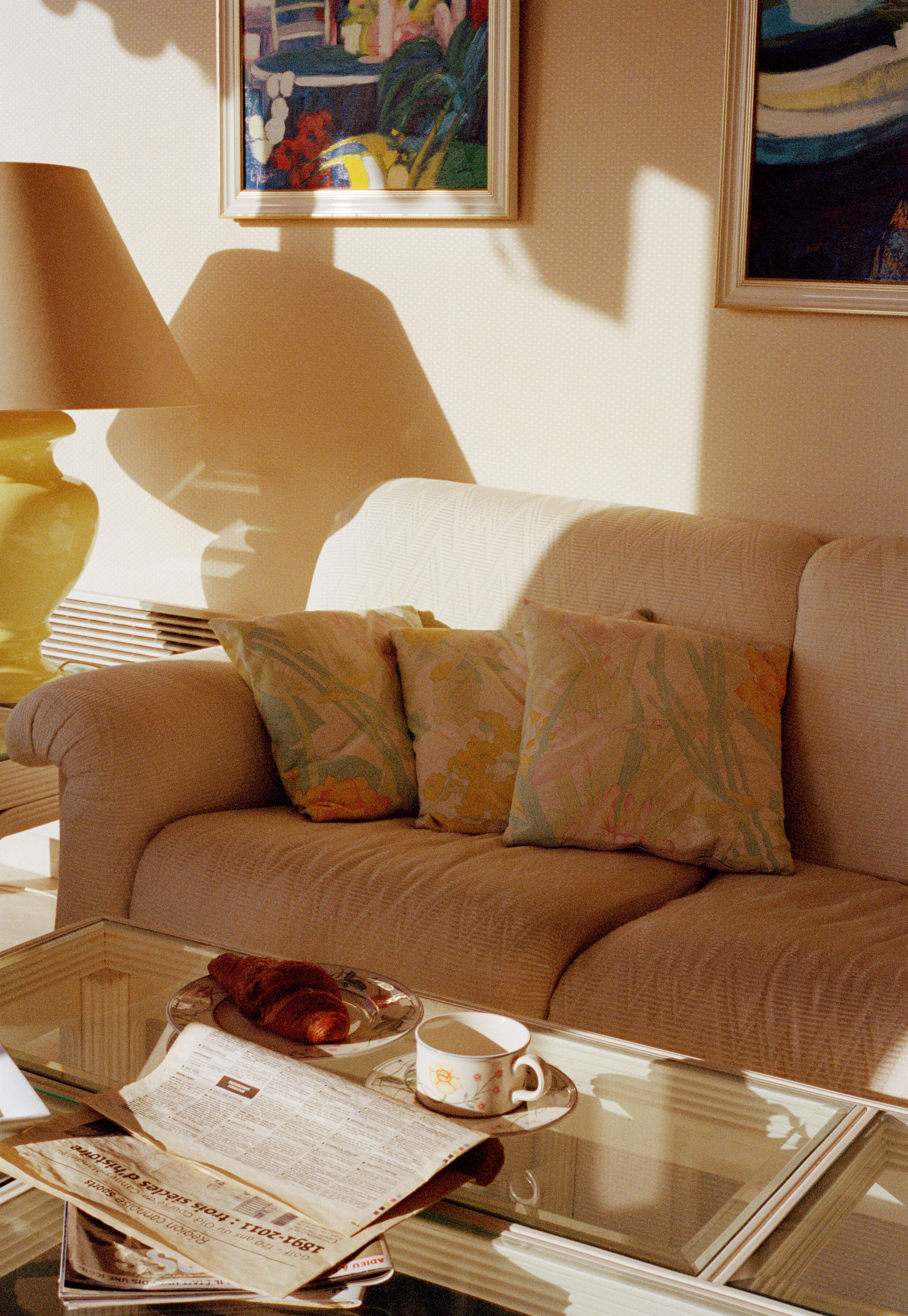 A living room with a beige couch, floral pillows, a glass coffee table with newspapers, a croissant, and a teacup, a yellow table lamp, and framed colorful artwork on the wall.