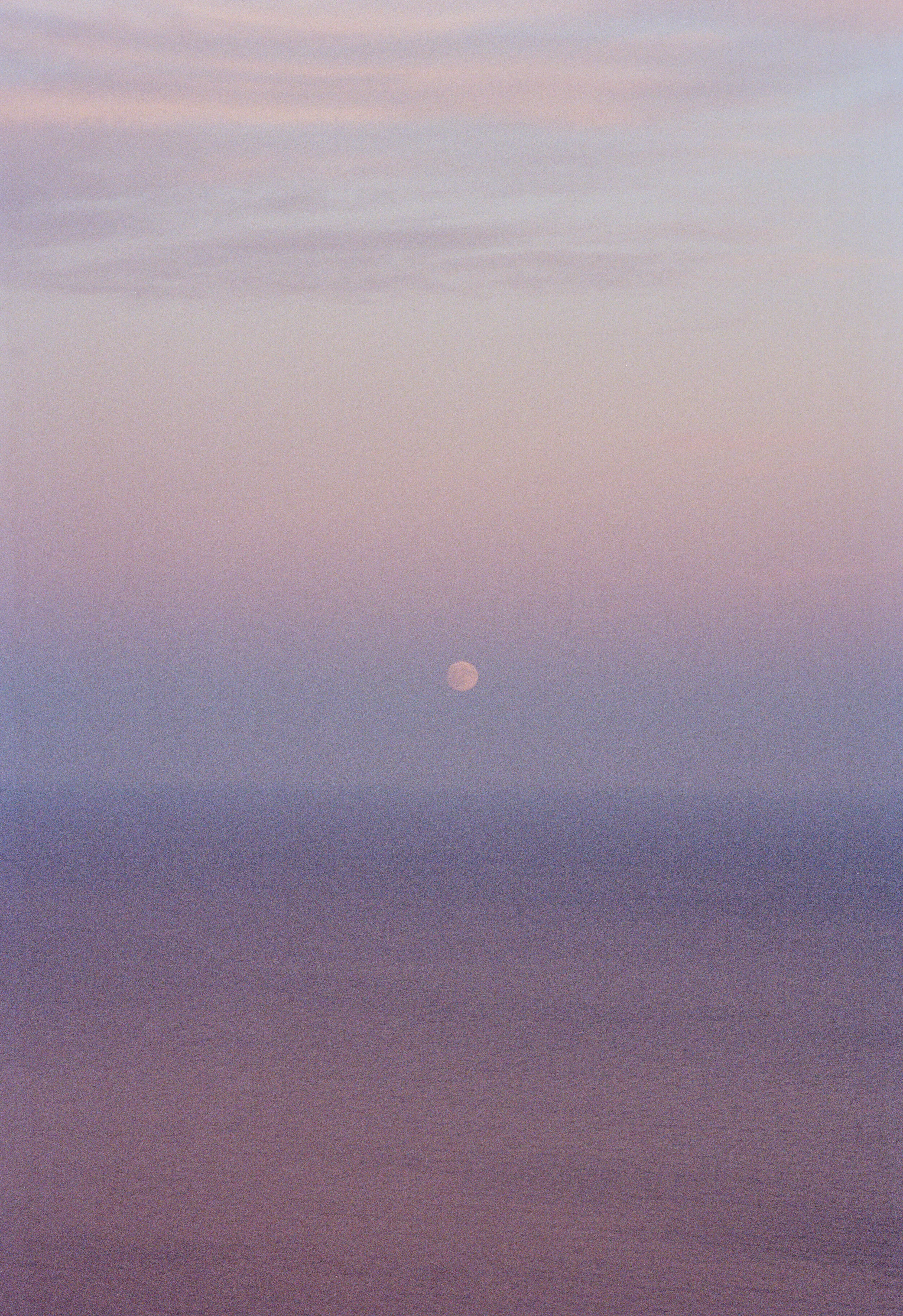 A photograph of the moon over the ocean at sunset, with soft pastel colors in the sky.